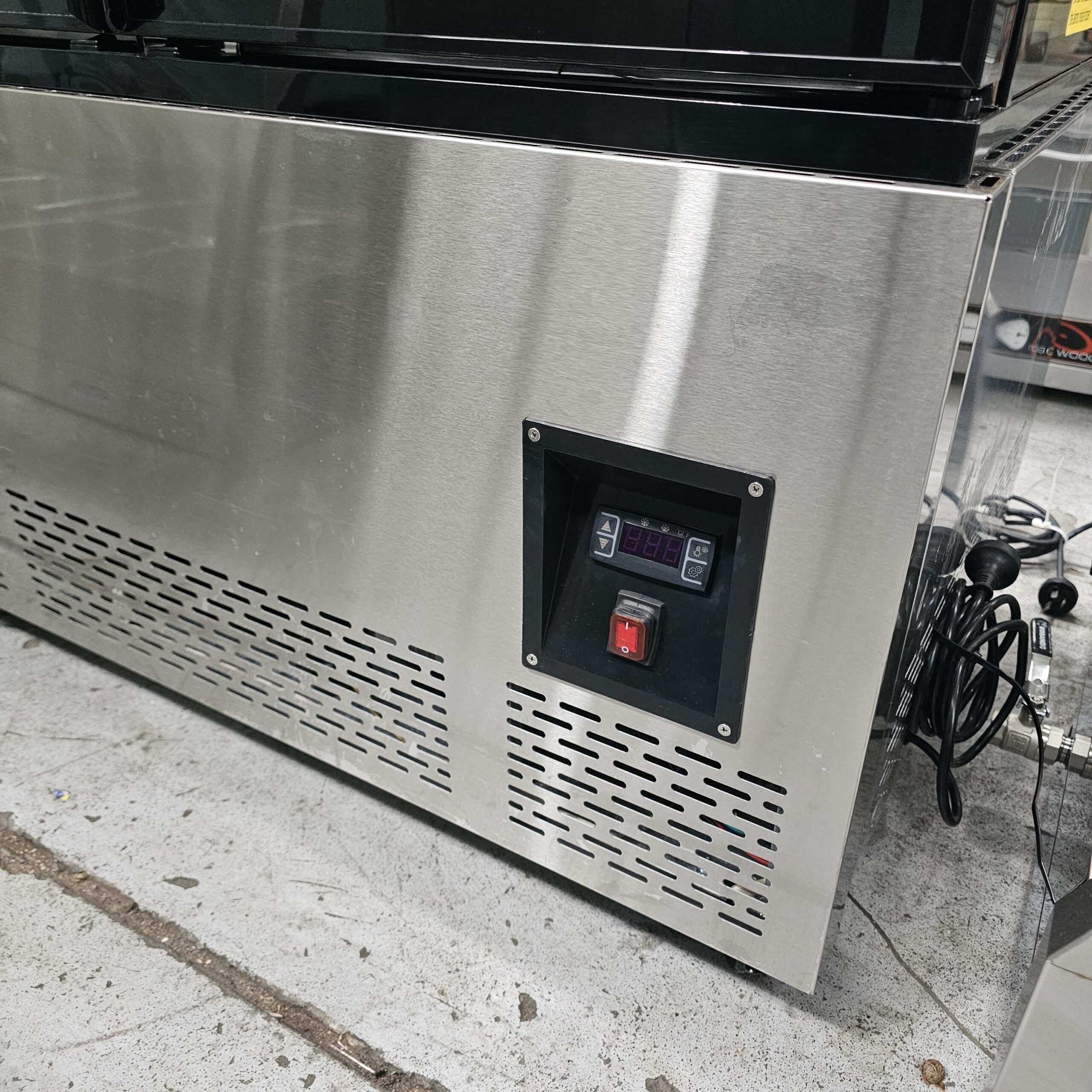 A close-up of a stainless steel appliance with a vented panel and a digital control display, red switch, and a visible power cord, situated on a speckled concrete floor. MRCE