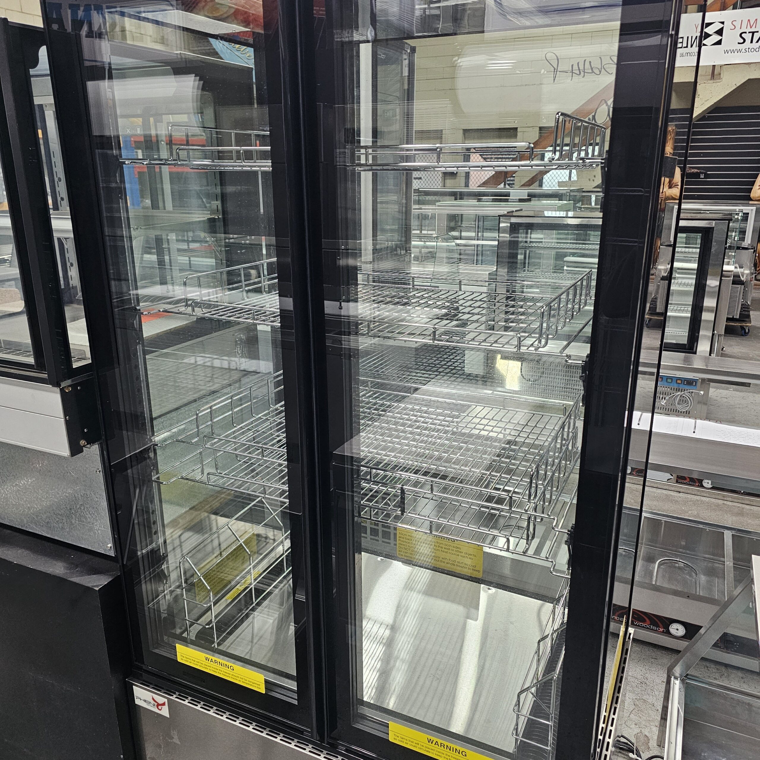 A commercial glass-door refrigerator with empty metal shelves, seen in what appears to be a store or supermarket, with reflections visible on the glass. MRCE