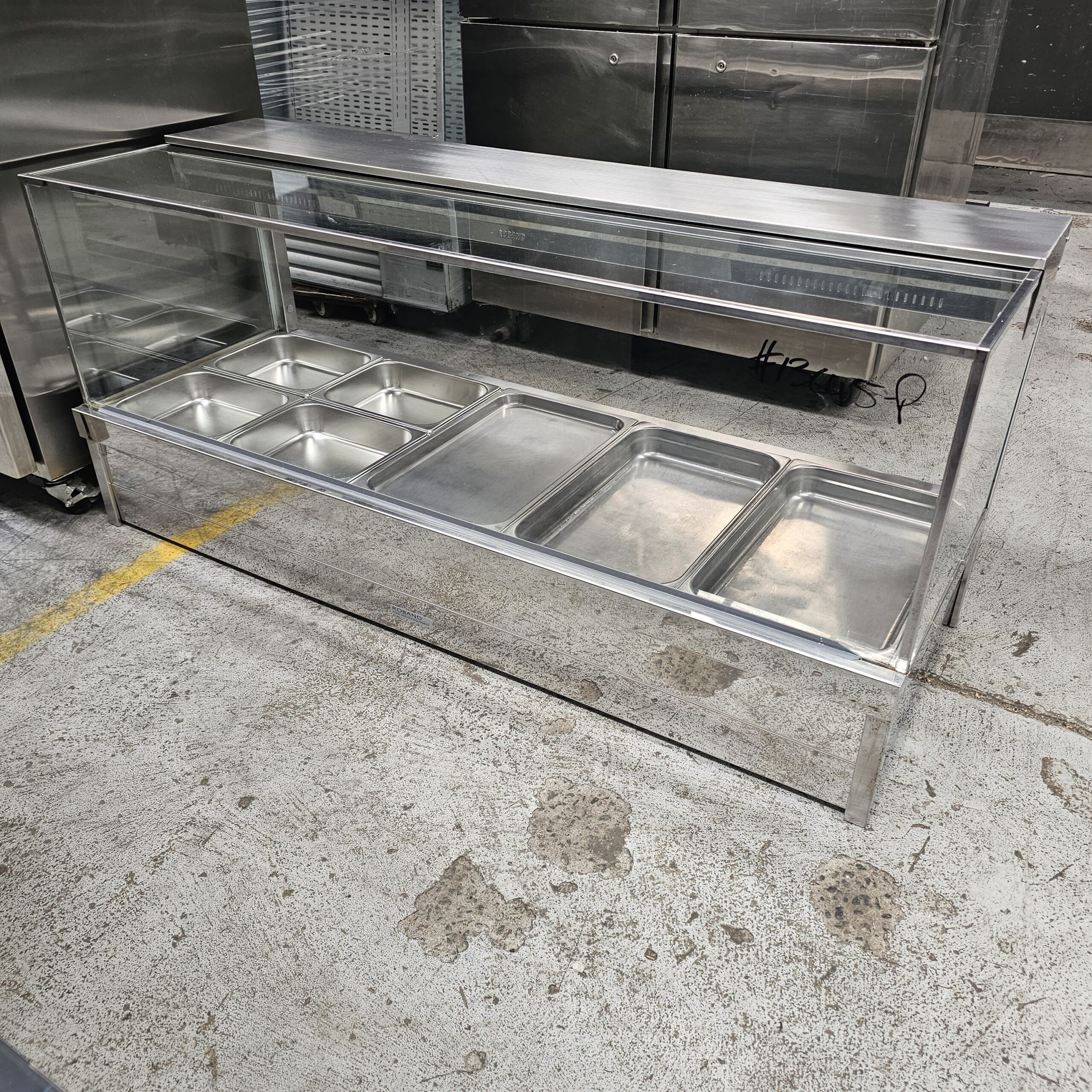 A stainless steel food service counter with a glass sneeze guard, featuring multiple empty metal trays, stands on a concrete floor in an industrial kitchen setting. MRCE