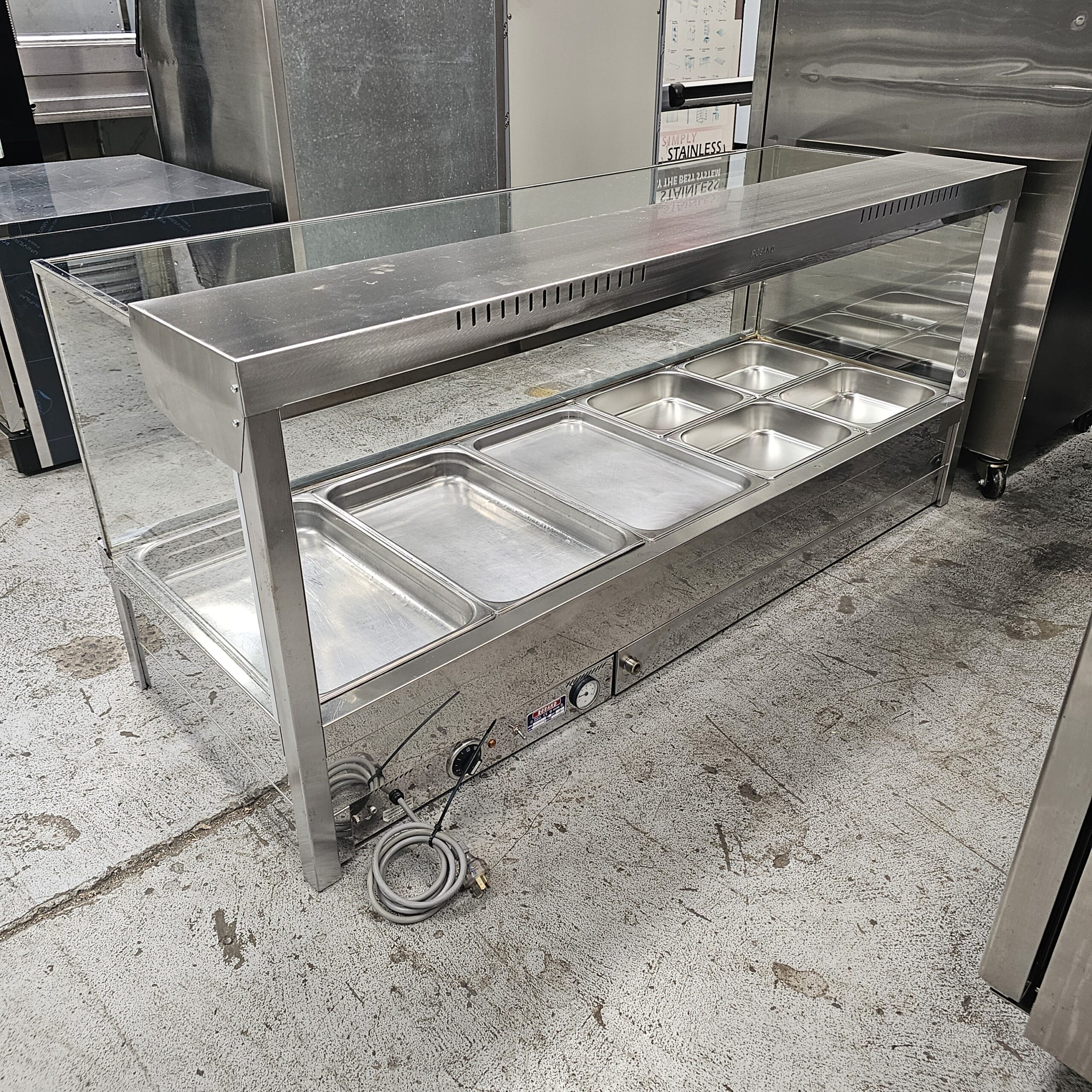 A stainless steel commercial food warmer with six empty food pans, glass sneeze guard, and control knobs beneath, placed on a gray, textured floor in a kitchen or cafeteria setting. MRCE