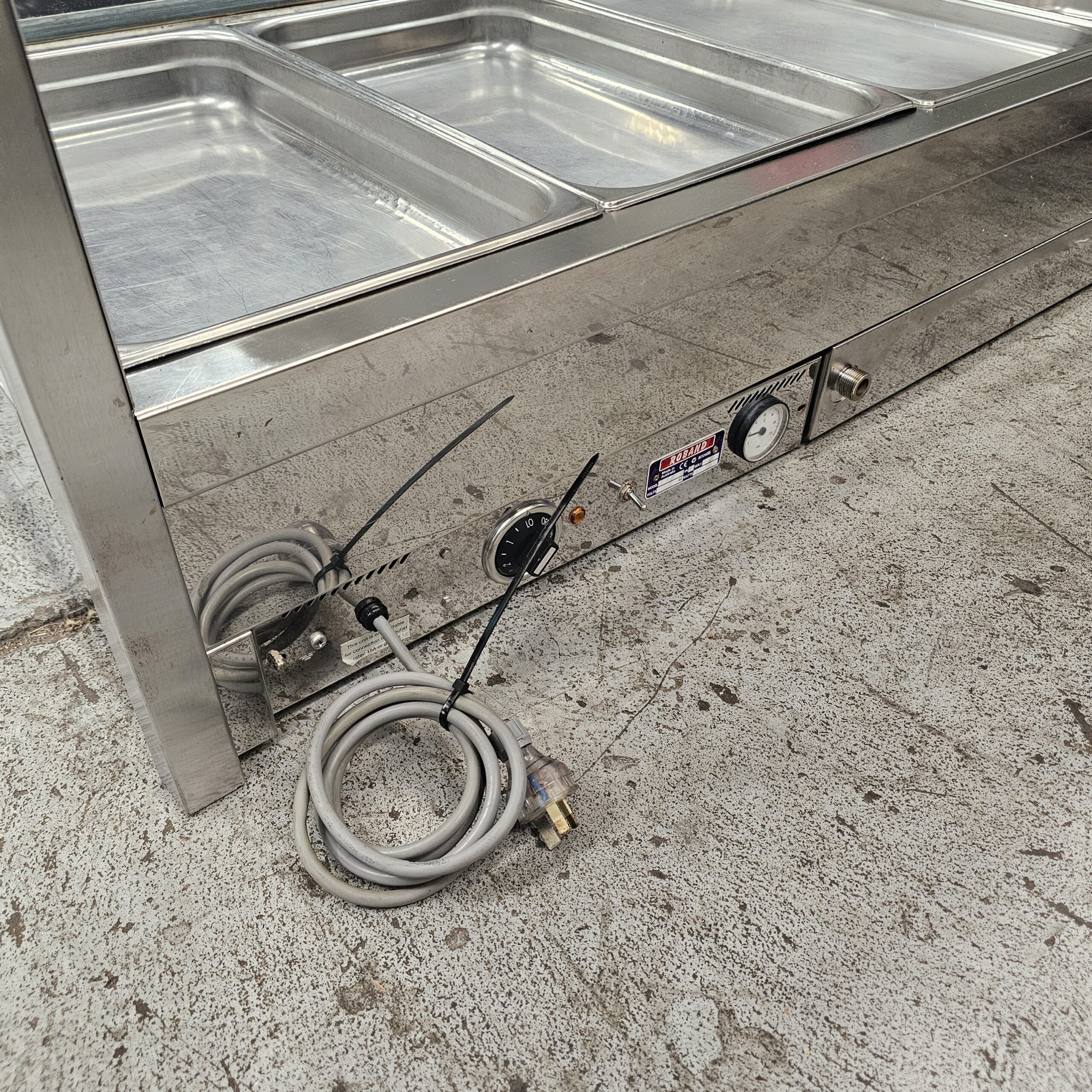 A stainless steel food warmer with empty trays is shown on a concrete floor. A coiled power cord is tied with a zip tie near a control dial and a temperature gauge on the side of the warmer. MRCE