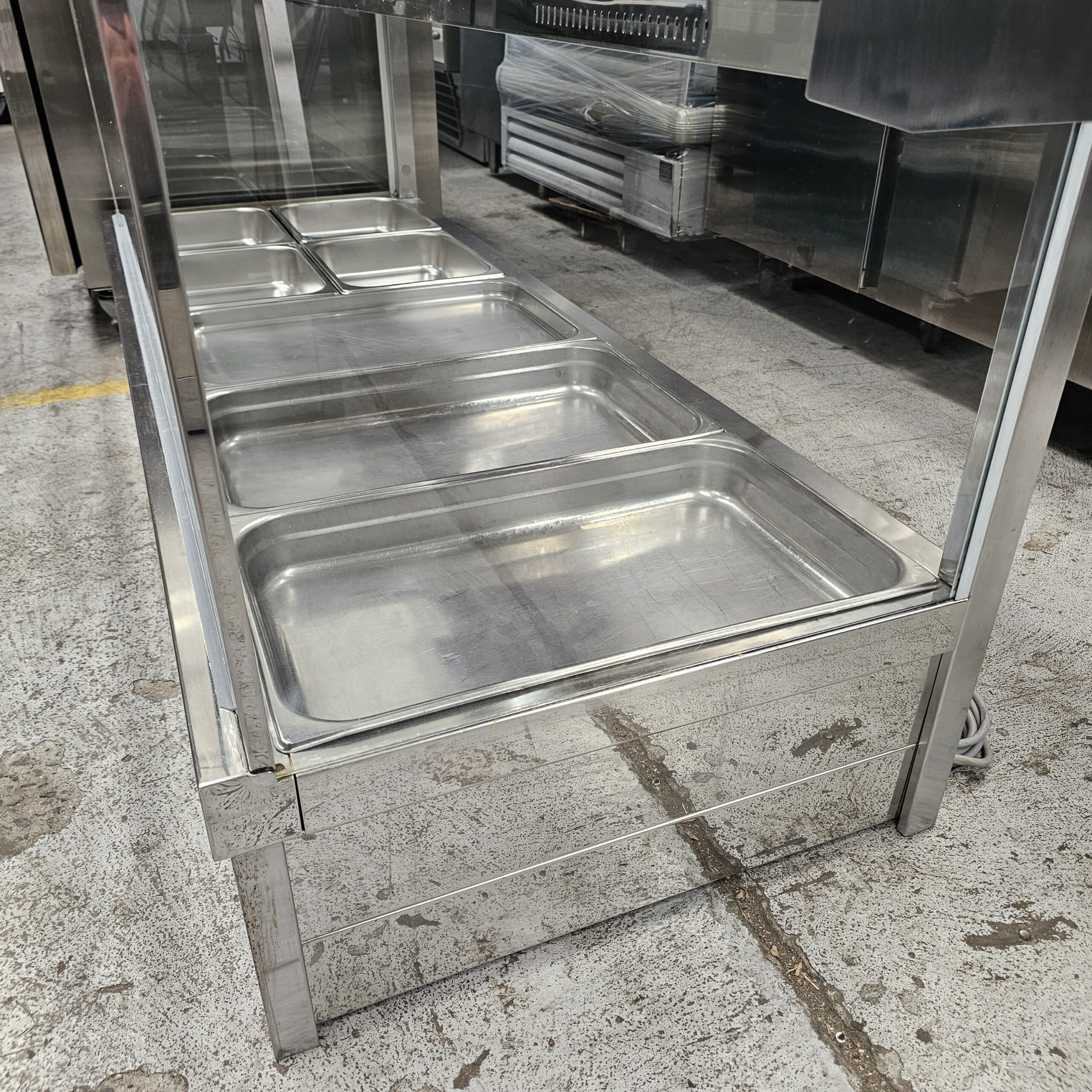 A stainless steel commercial food service counter with several empty metal trays, set on a concrete floor in an industrial or kitchen setting. MRCE