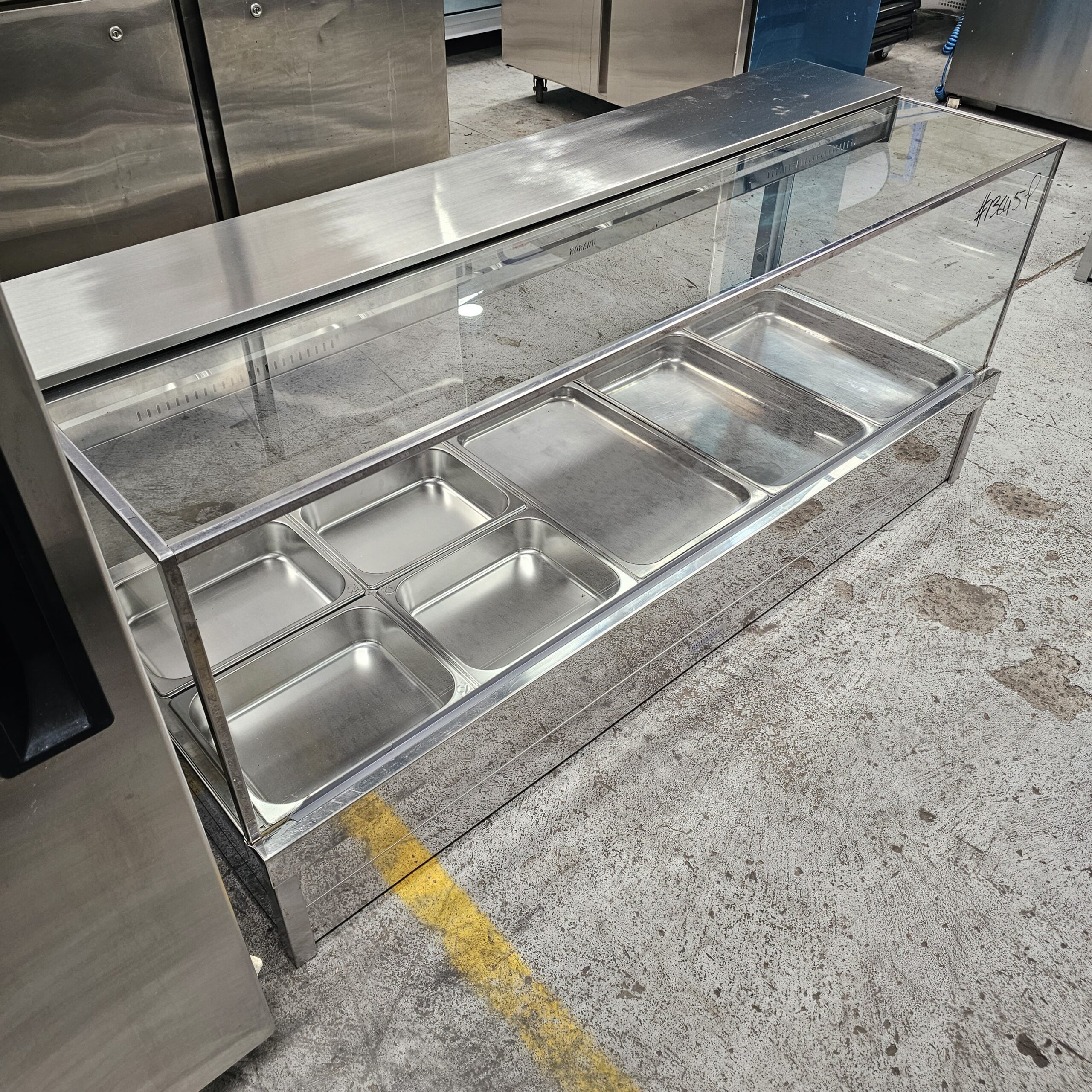 A stainless steel food service counter with two shelves, each holding several empty metal trays, stands on a concrete floor with a yellow line nearby. The counter has a glass sneeze guard on the top front. MRCE