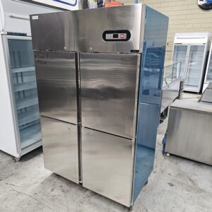 A large commercial stainless steel refrigerator with four doors stands on a warehouse floor, surrounded by other industrial kitchen appliances and equipment. MRCE