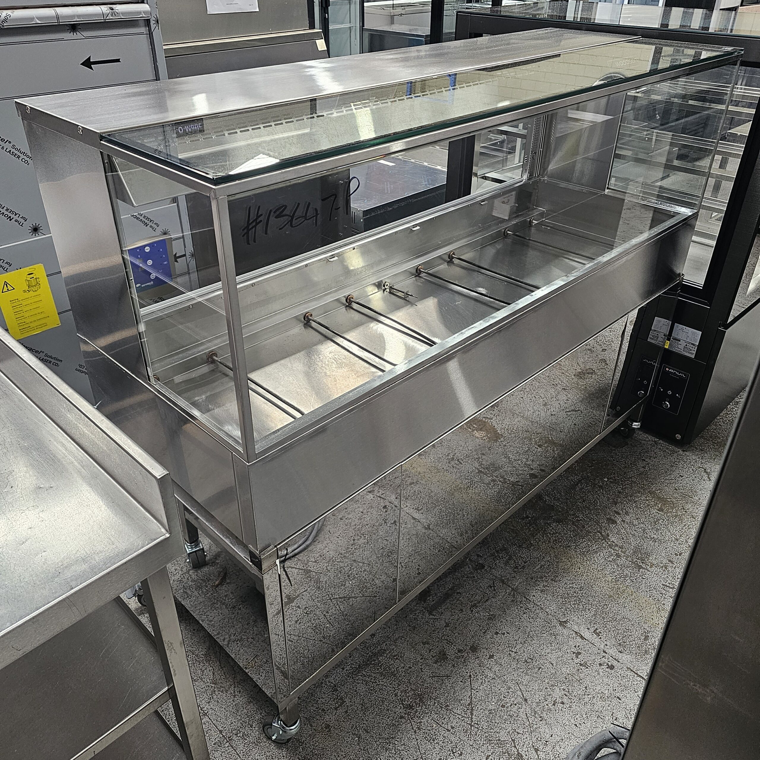 A stainless steel commercial food display case with a glass top and sides, featuring multiple food tray slots, stands on wheels in a showroom with other kitchen equipment. MRCE
