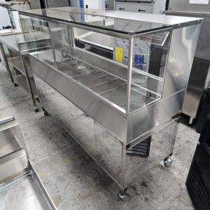 A stainless steel and glass display case with wheels, designed for food service, stands empty on a concrete floor in a commercial kitchen or equipment showroom. MRCE
