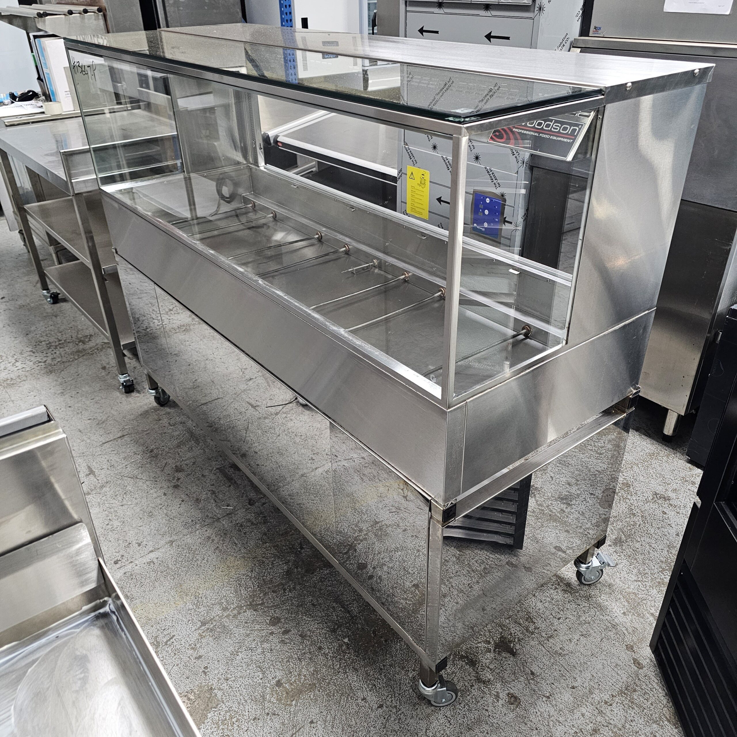 A stainless steel and glass display case with wheels, designed for food service, stands empty on a concrete floor in a commercial kitchen or equipment showroom. MRCE