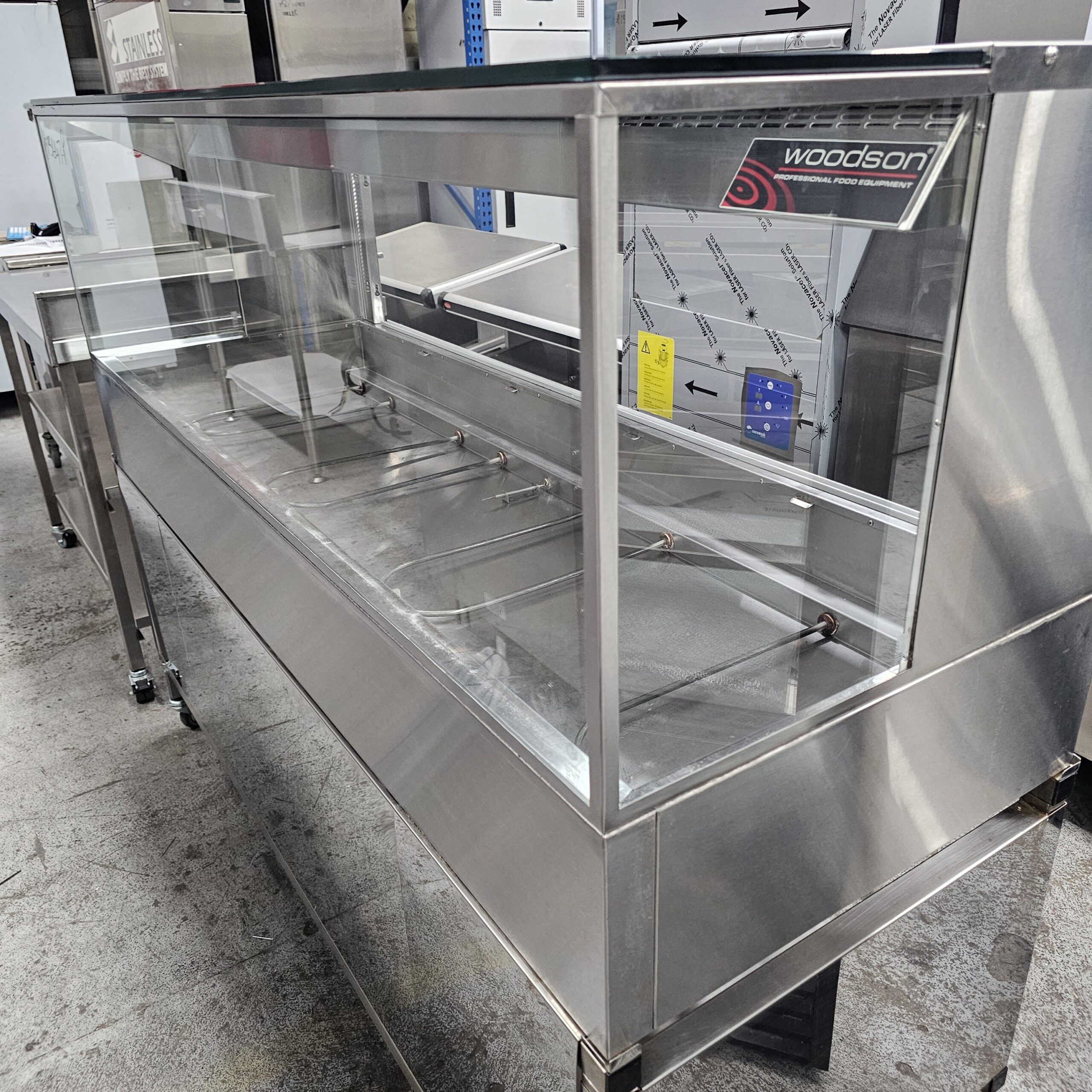 A large empty stainless steel commercial food display cabinet with a glass front and top, located in what appears to be a storage or commercial kitchen area. MRCE