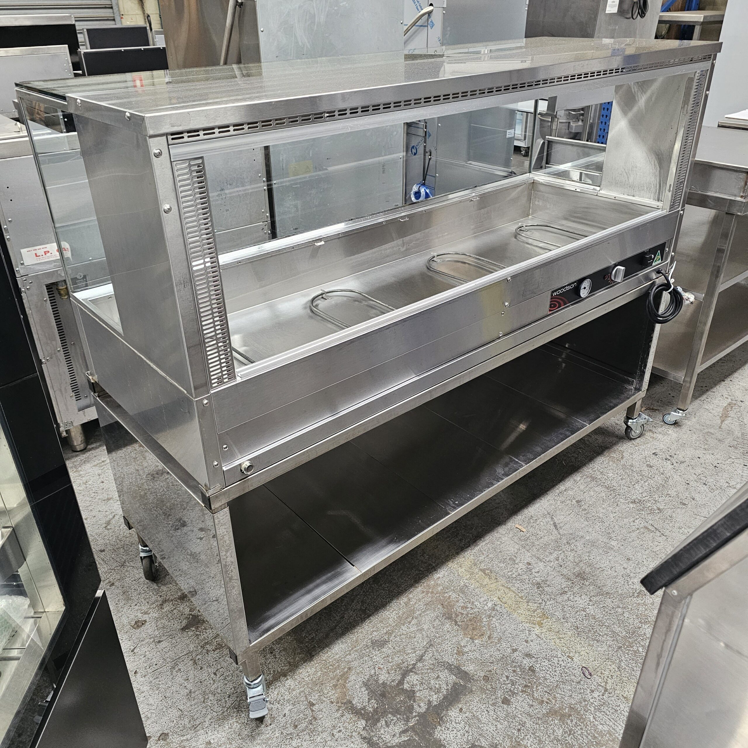 A stainless steel commercial food warmer with glass sides, three covered food compartments, control knobs, and a storage shelf underneath, standing on wheels in an industrial kitchen setting. MRCE