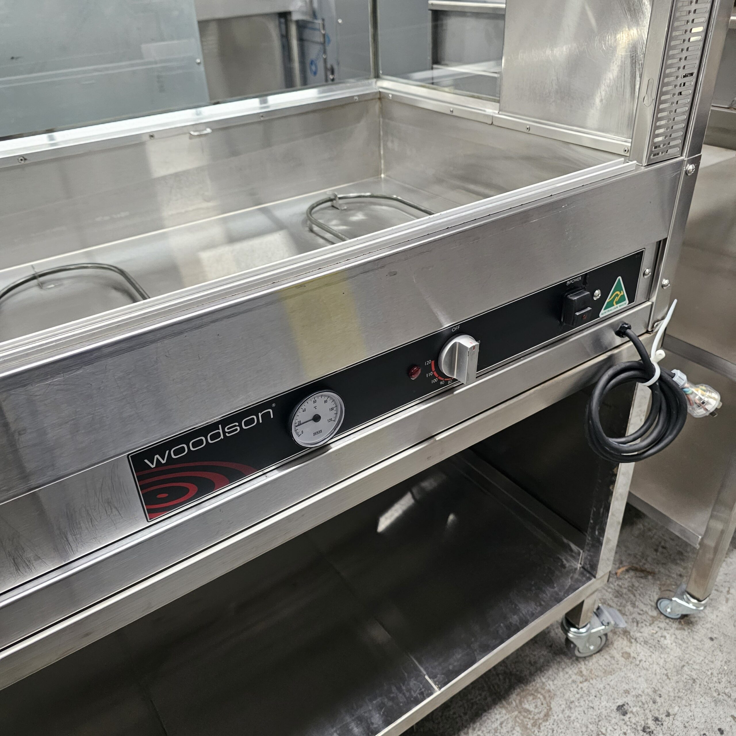 A stainless steel commercial food warmer by Woodson with a temperature dial, power indicator lights, and a coiled power cord, standing on wheels in a kitchen or food service area. MRCE