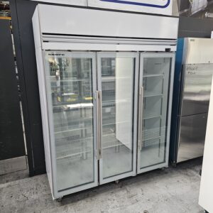 A large commercial refrigerator with three transparent glass doors, empty white shelves inside, and metal handles, situated on a warehouse floor. MRCE