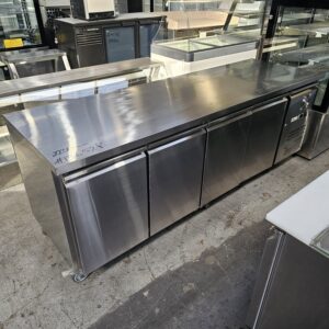 A large stainless steel commercial refrigerator with three doors, set in an industrial kitchen or warehouse with other appliances visible in the background. MRCE