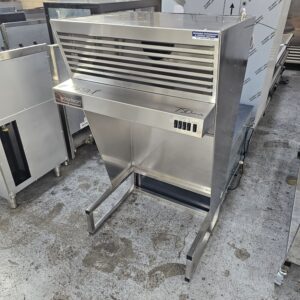 A large stainless steel commercial kitchen appliance with vented front, control switches, and an open lower frame, standing on a concrete floor among other metal equipment. MRCE