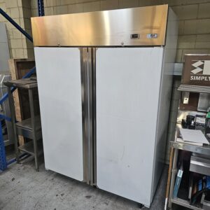 A large, commercial double-door stainless steel refrigerator stands in an industrial setting by metal shelves and various equipment, with a beige and silver top panel and a digital display. MRCE