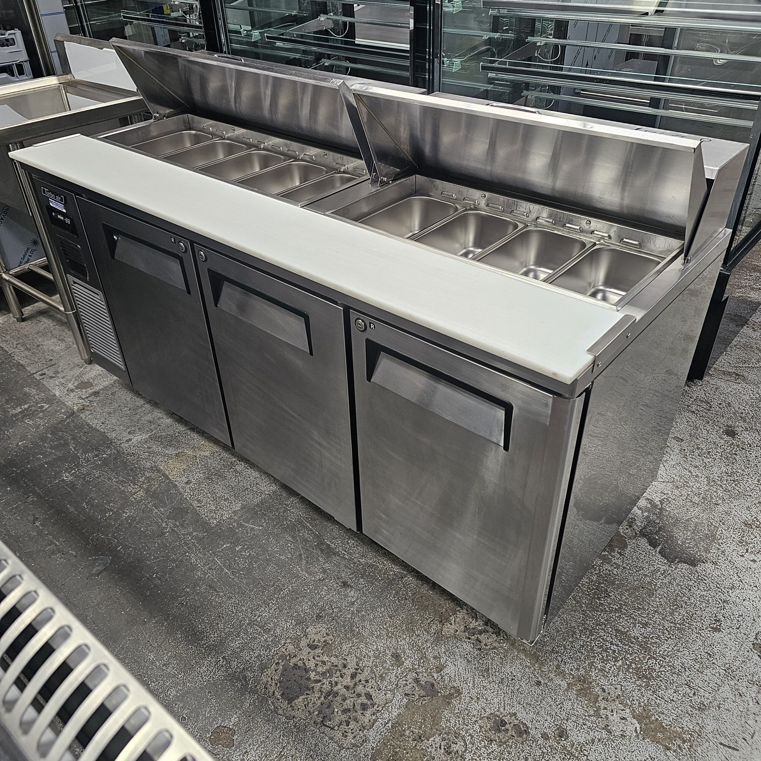 A commercial stainless steel refrigerated prep table with a white cutting surface and several covered food pans, situated in a kitchen or industrial setting. MRCE