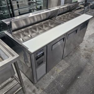 A stainless steel commercial prep table with an open refrigerated top holding multiple empty metal food pans, a white cutting board on the front, and two closed storage compartments below. MRCE