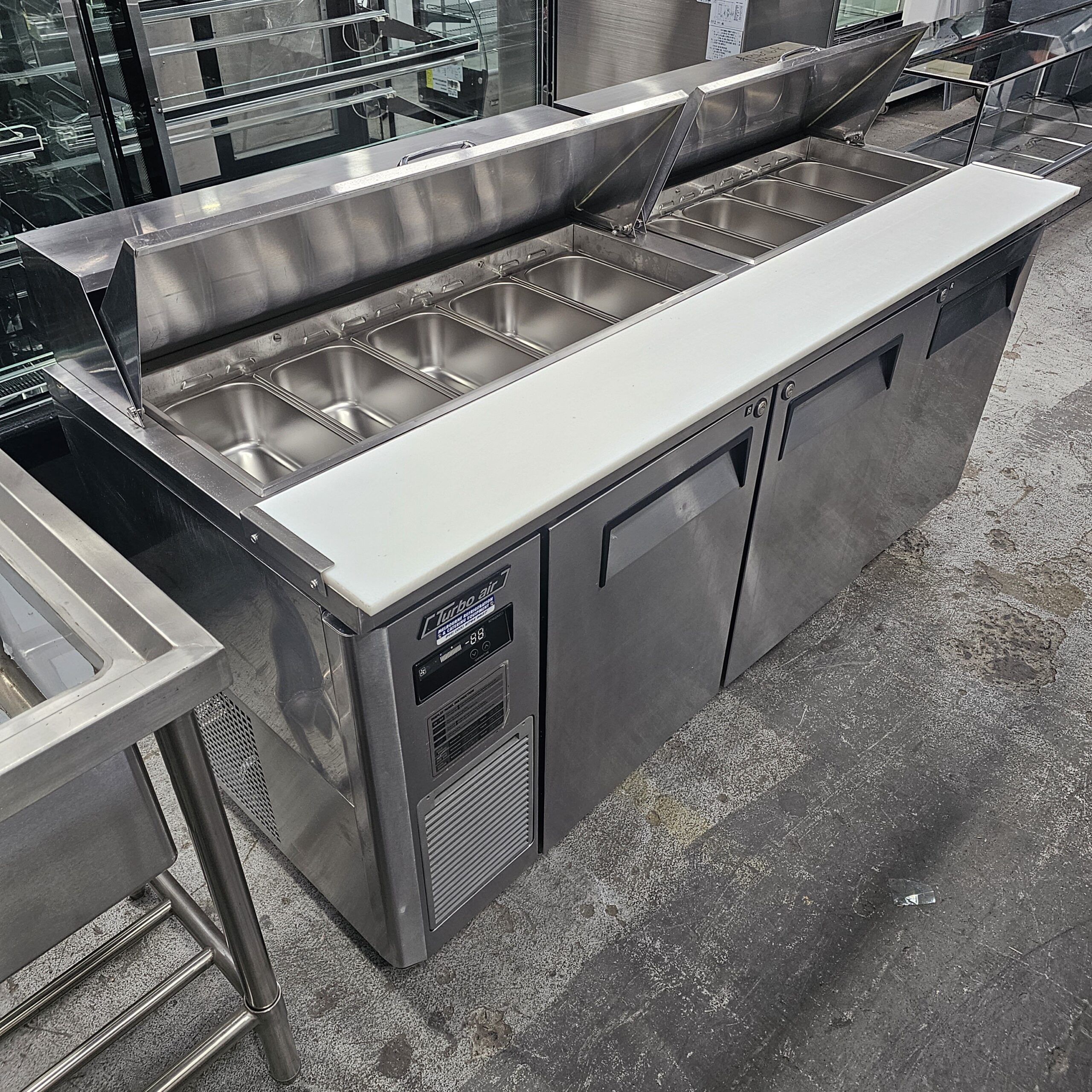 A stainless steel commercial prep table with an open refrigerated top holding multiple empty metal food pans, a white cutting board on the front, and two closed storage compartments below. MRCE
