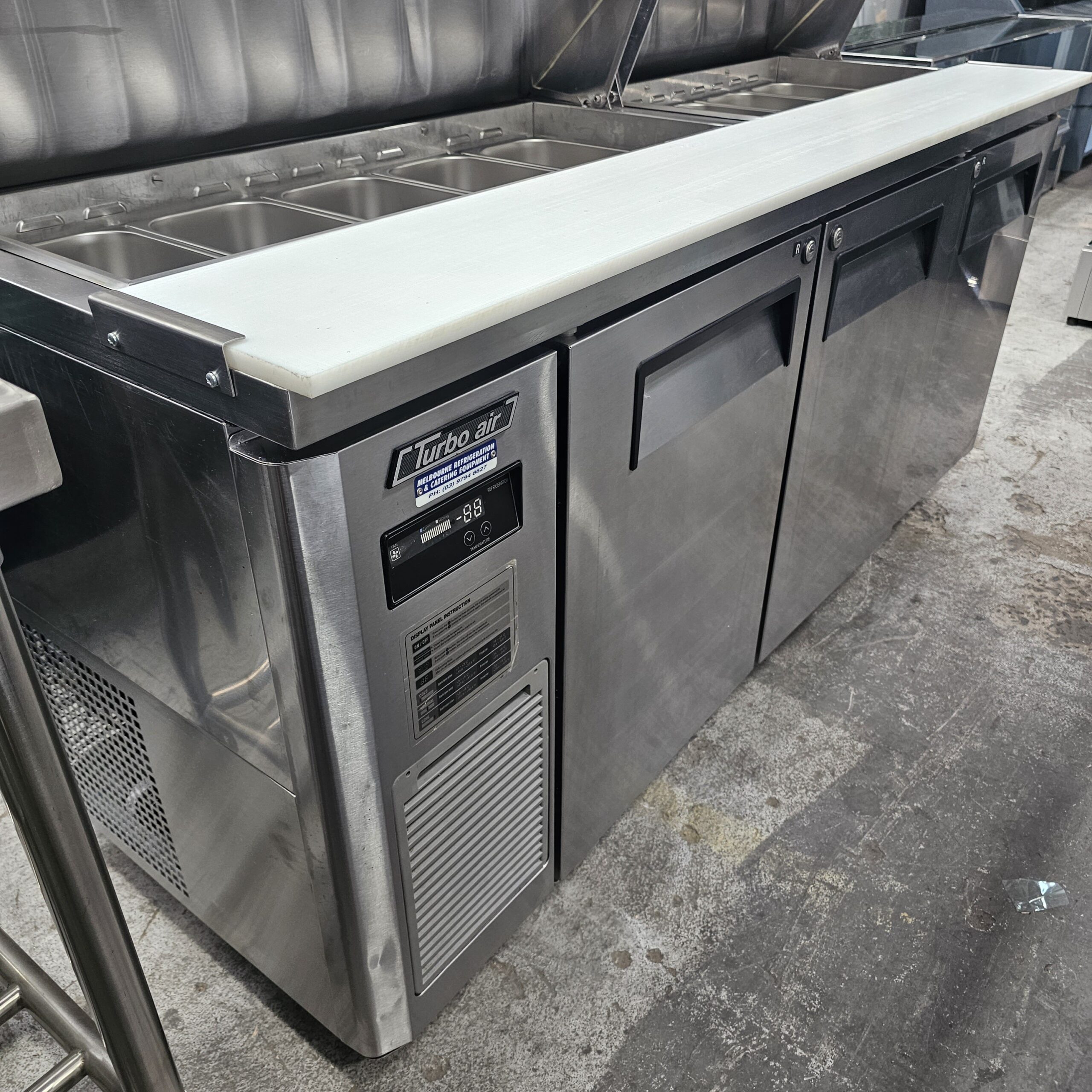 A stainless steel commercial refrigerator with a white countertop, digital temperature display, and multiple food storage compartments on top, situated in a professional kitchen. MRCE