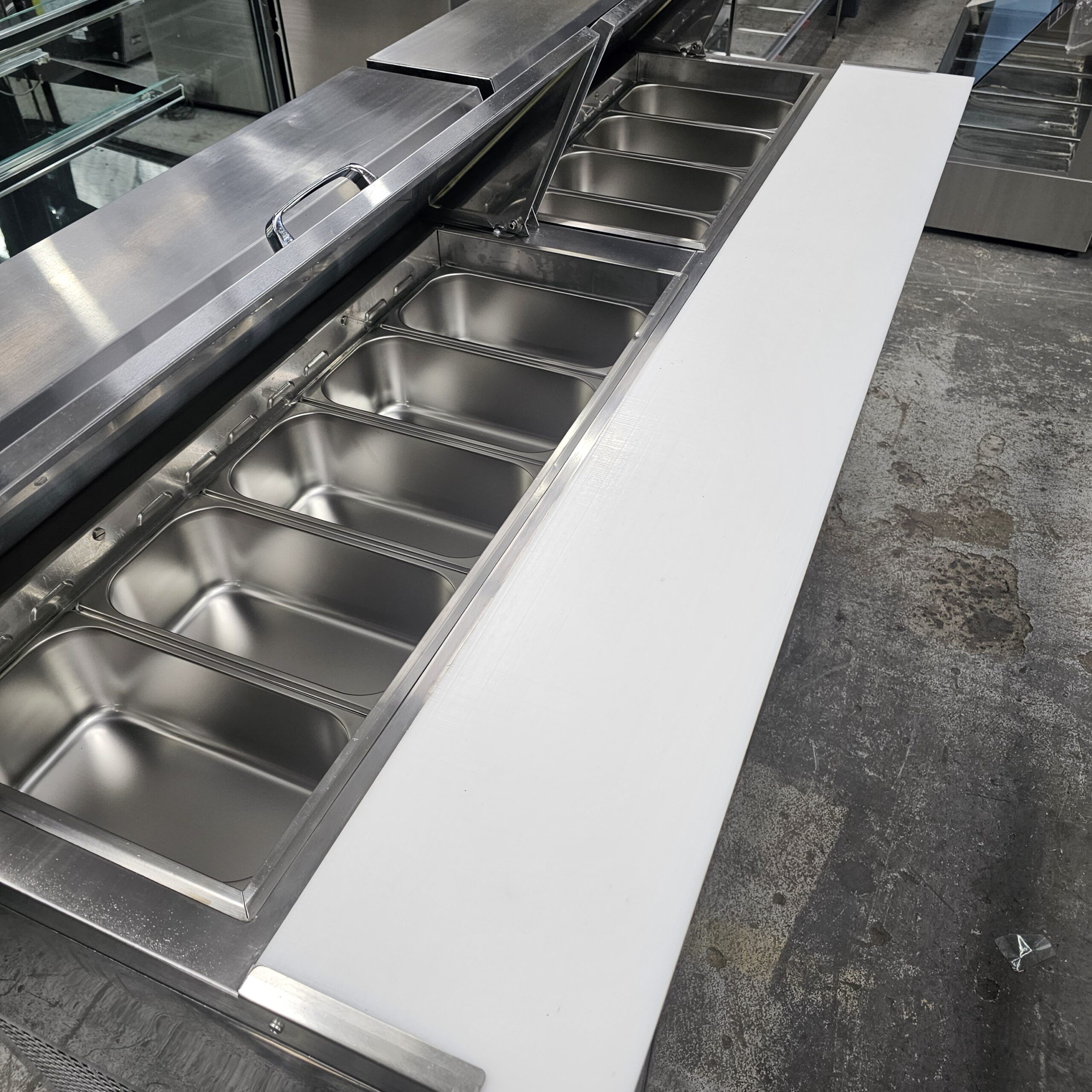 A commercial stainless steel prep table with multiple empty food pans and a large white cutting board attached in front, seen in an industrial kitchen setting. MRCE