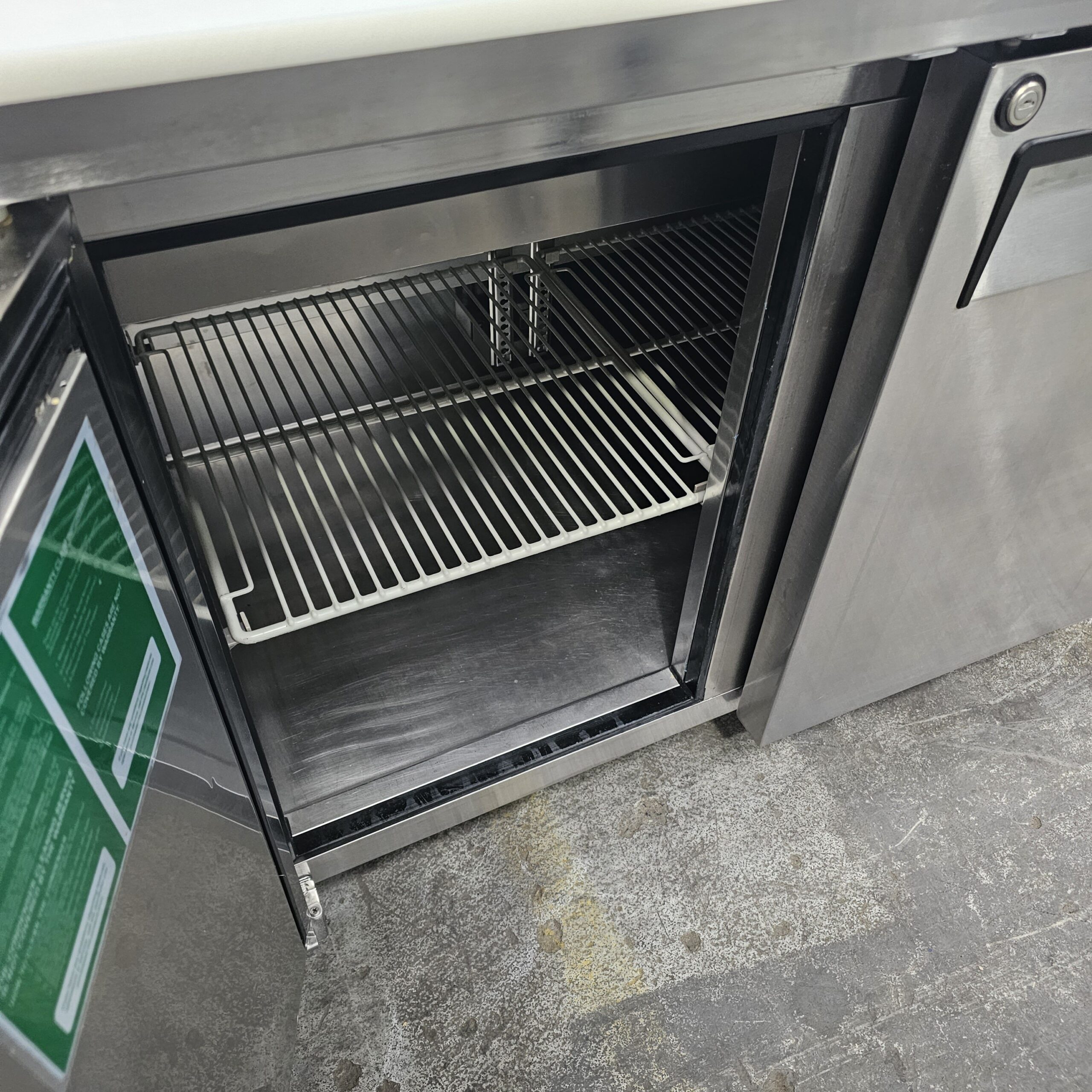 An empty stainless steel commercial refrigerator with its door open, showing two metal shelves inside. The fridge is set on a gray, textured floor. MRCE