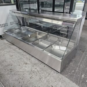 A stainless steel commercial food display counter with a glass sneeze guard, featuring six empty metal food trays, situated indoors on a concrete floor. MRCE
