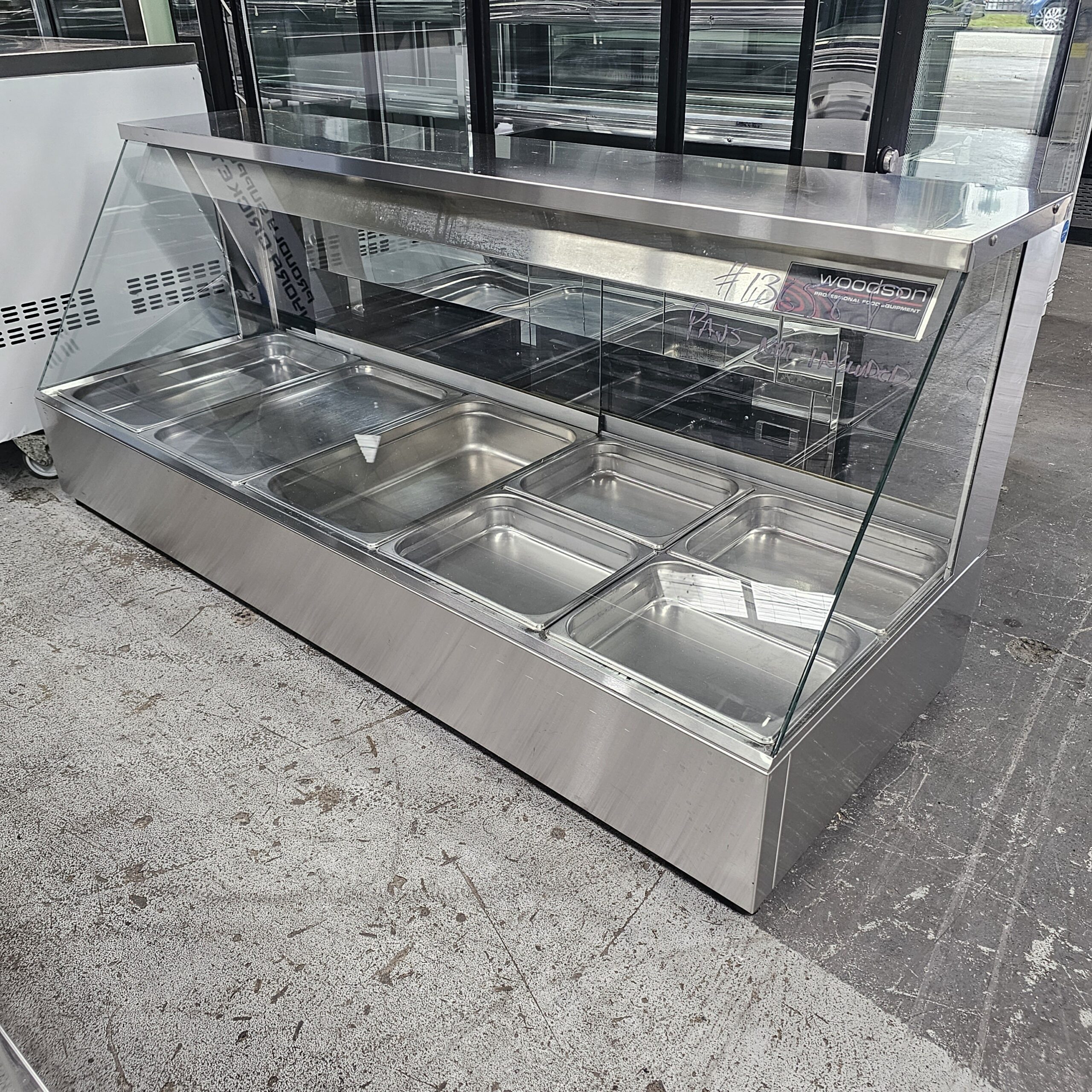 A stainless steel commercial food display counter with a glass sneeze guard, featuring six empty metal food trays, situated indoors on a concrete floor. MRCE