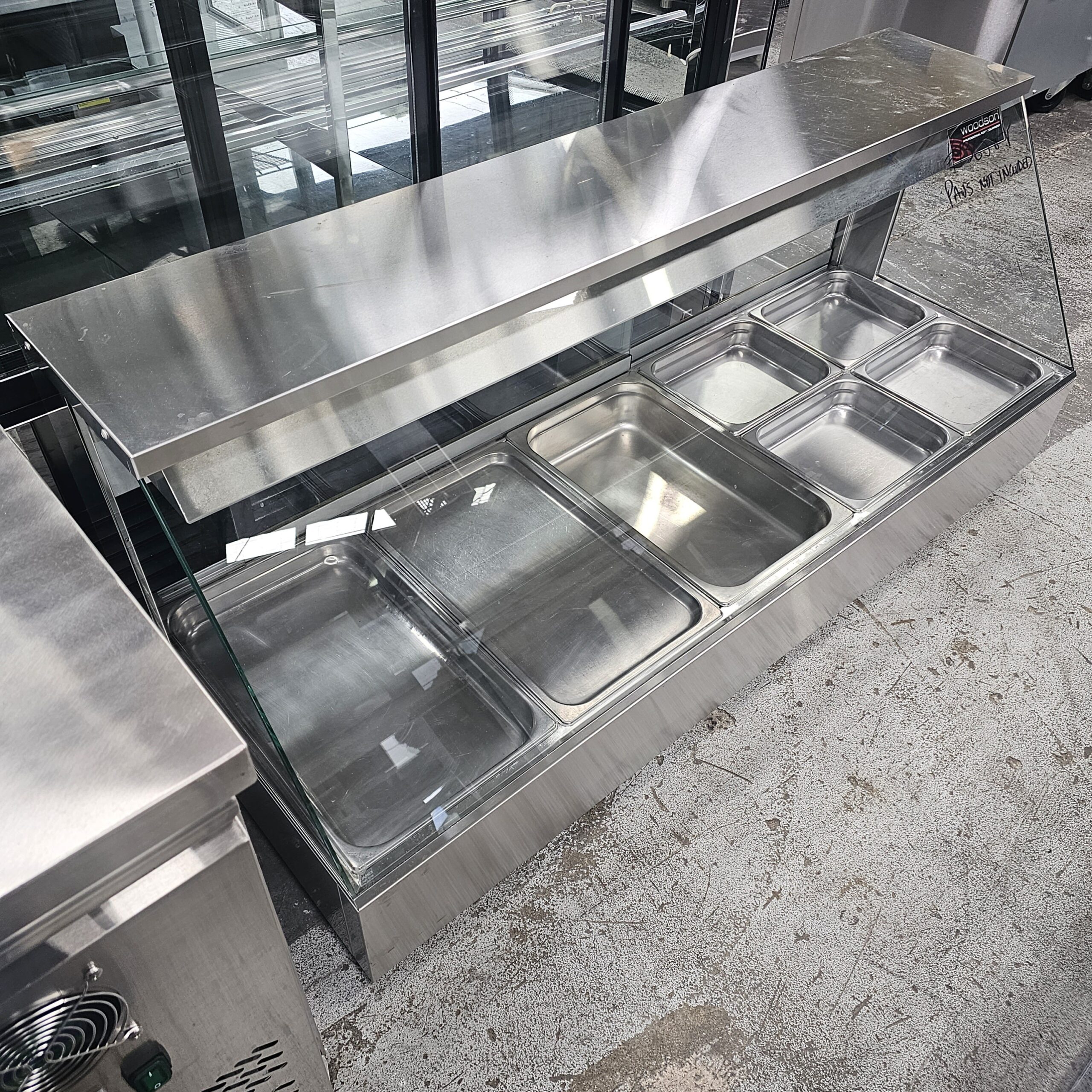 A stainless steel commercial food display case with a glass front, featuring eight empty metal food pans, placed on a light-colored concrete floor in an indoor setting. MRCE
