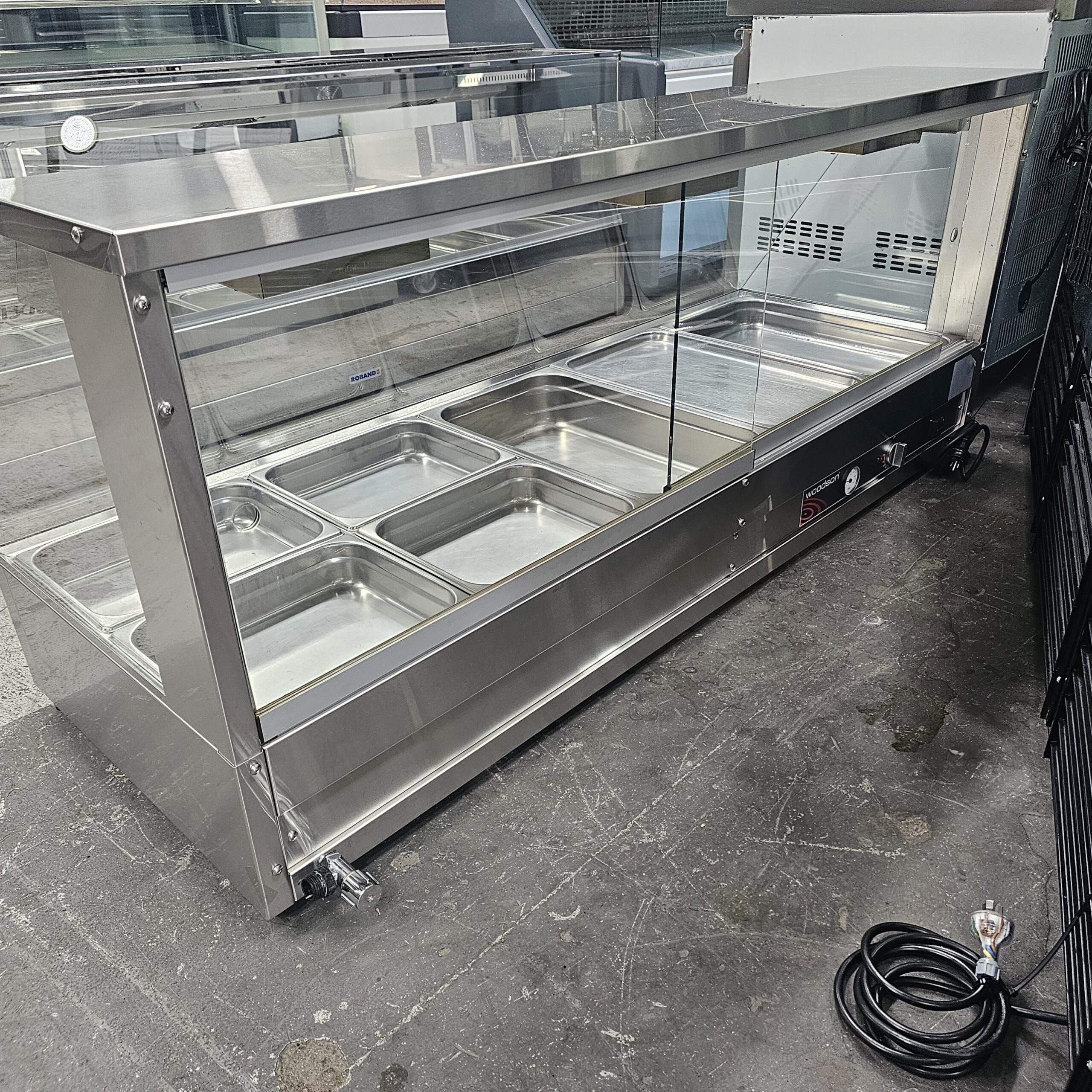 A stainless steel food display counter with six empty metal trays, a glass front, and an electrical cord coiled on the floor nearby, is positioned on a concrete surface. MRCE