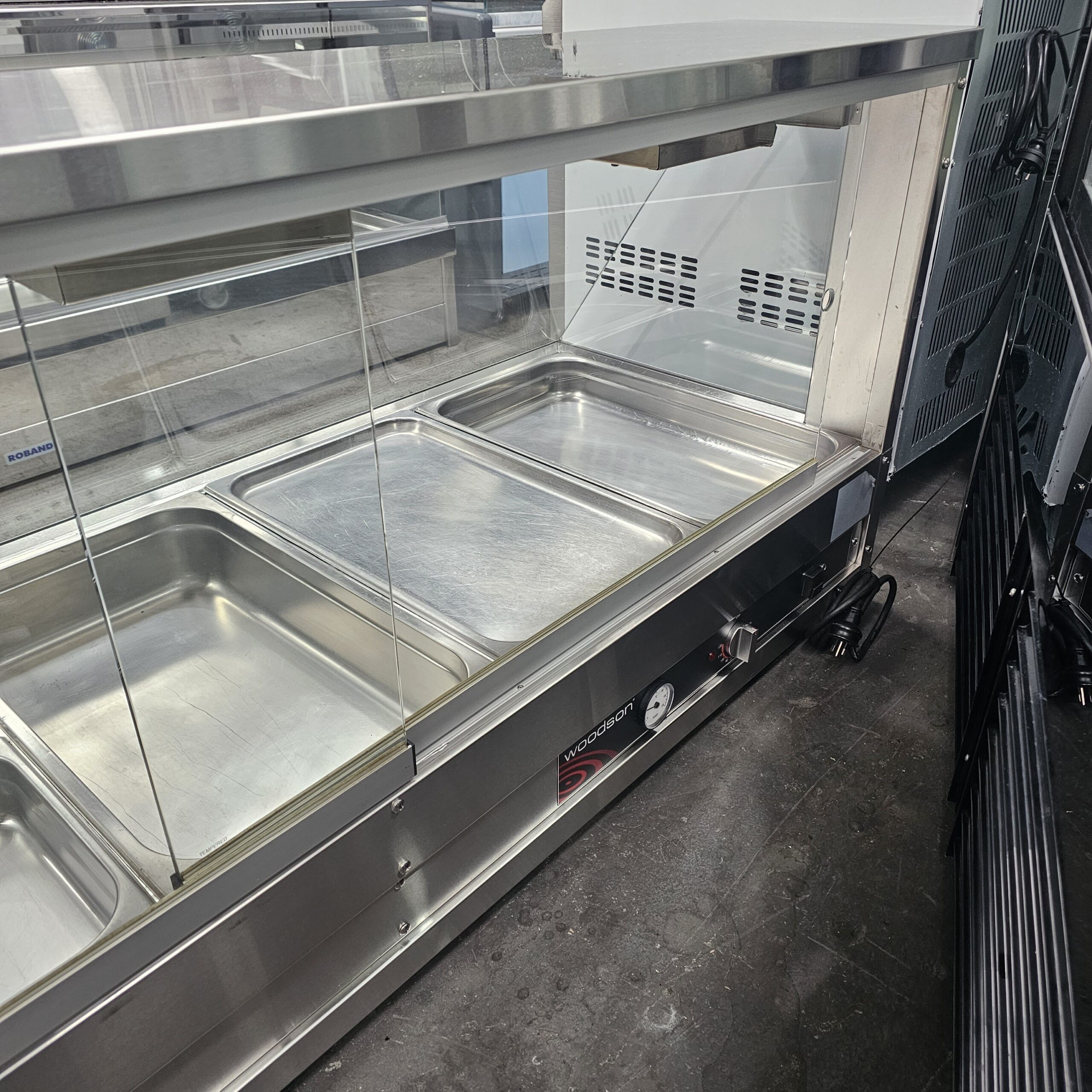 A stainless steel empty food service counter with several metal trays, covered by a glass sneeze guard, stands on a gray floor next to a metal rack. MRCE