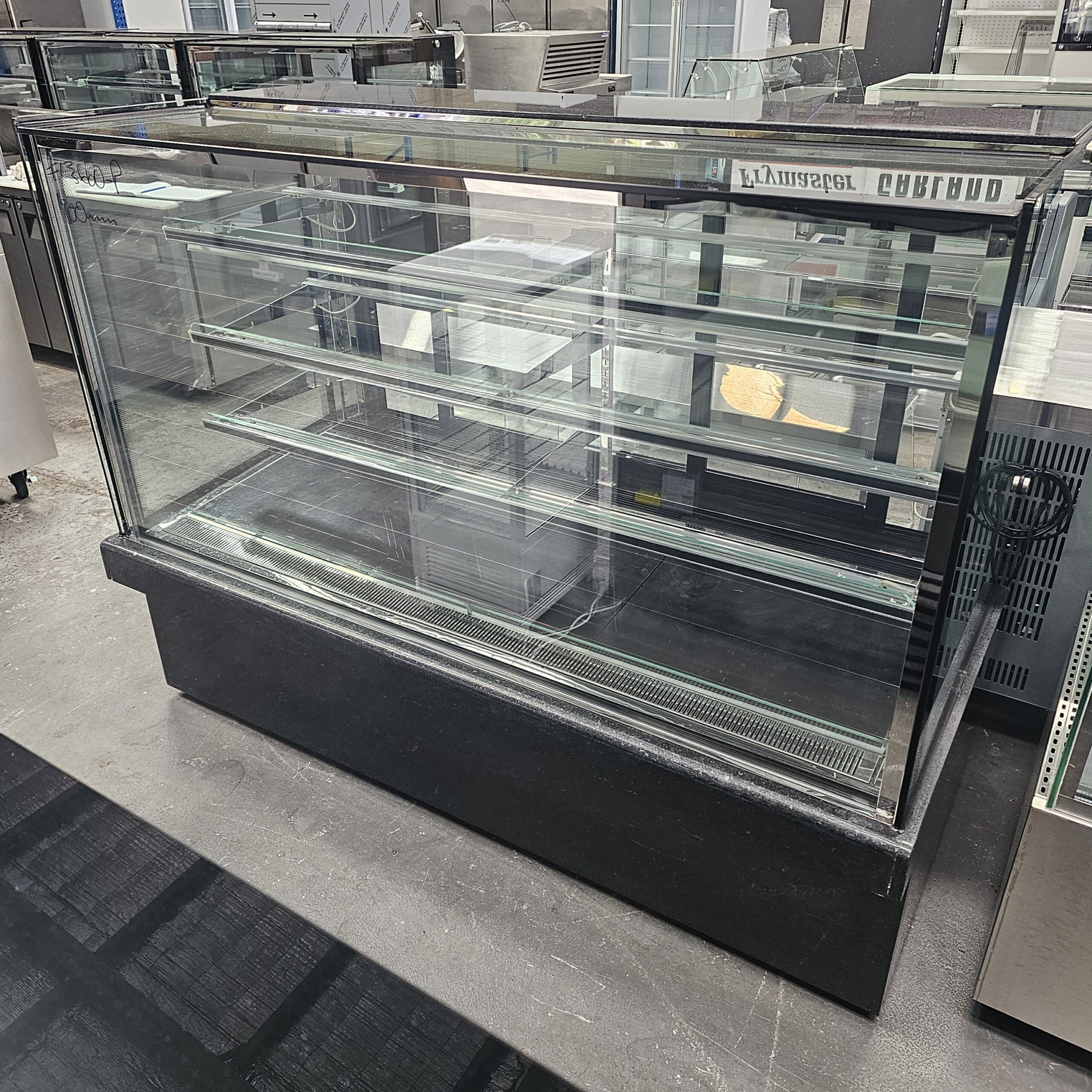 A large, empty glass display case with multiple shelves stands in a commercial setting, likely a bakery or café, surrounded by other metal and glass appliances. MRCE