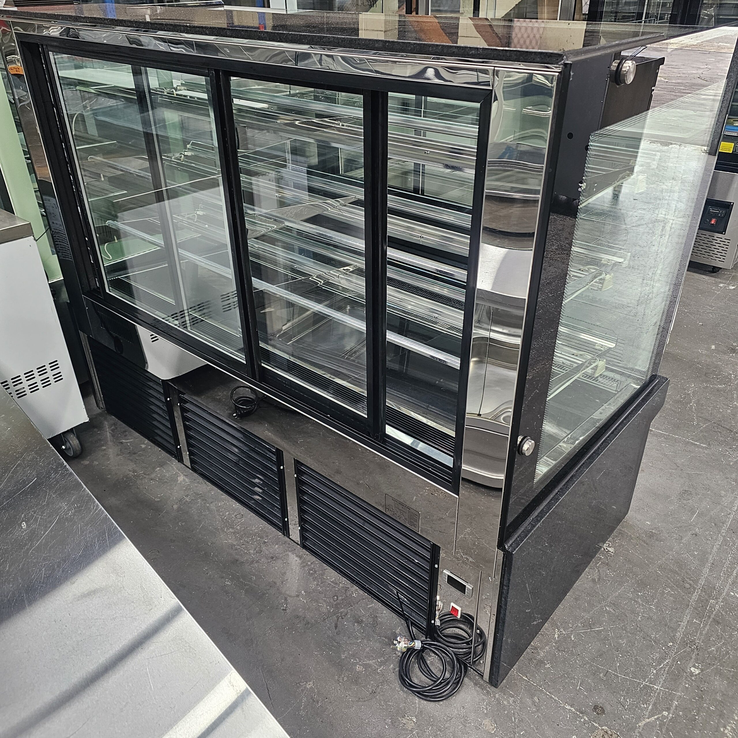 A glass display refrigerator with sliding doors, black frame, and multiple shelves, standing on a gray concrete floor in an industrial setting. Power cords are visible near the bottom. MRCE