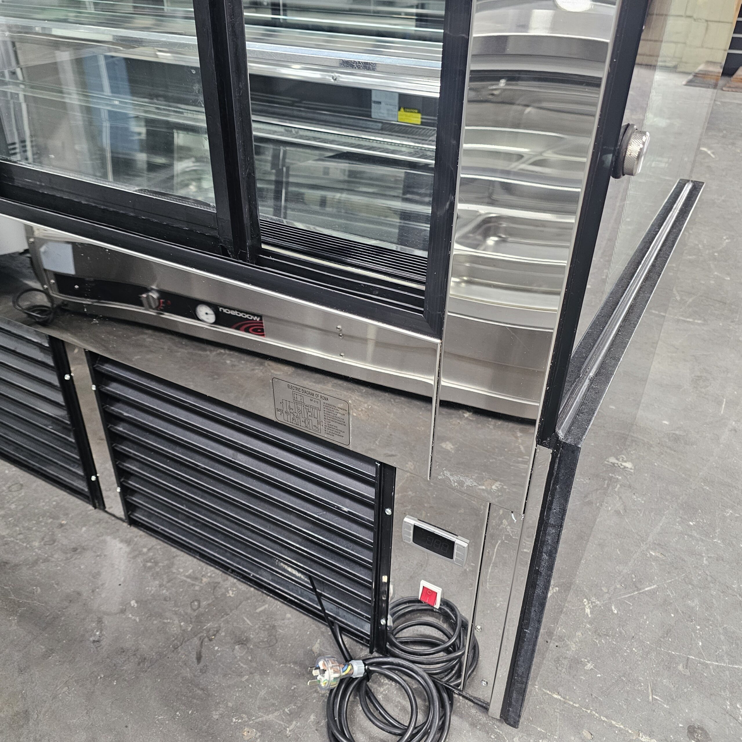 A commercial refrigerator with glass display doors stands open, showing its vented lower panel, power cord, and exposed controls, all placed on a gray concrete floor. MRCE