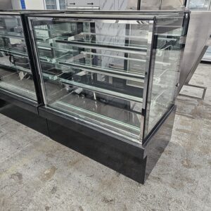 A large, empty glass display case with multiple shelves, standing on a black base in an industrial or commercial setting with a concrete floor. MRCE