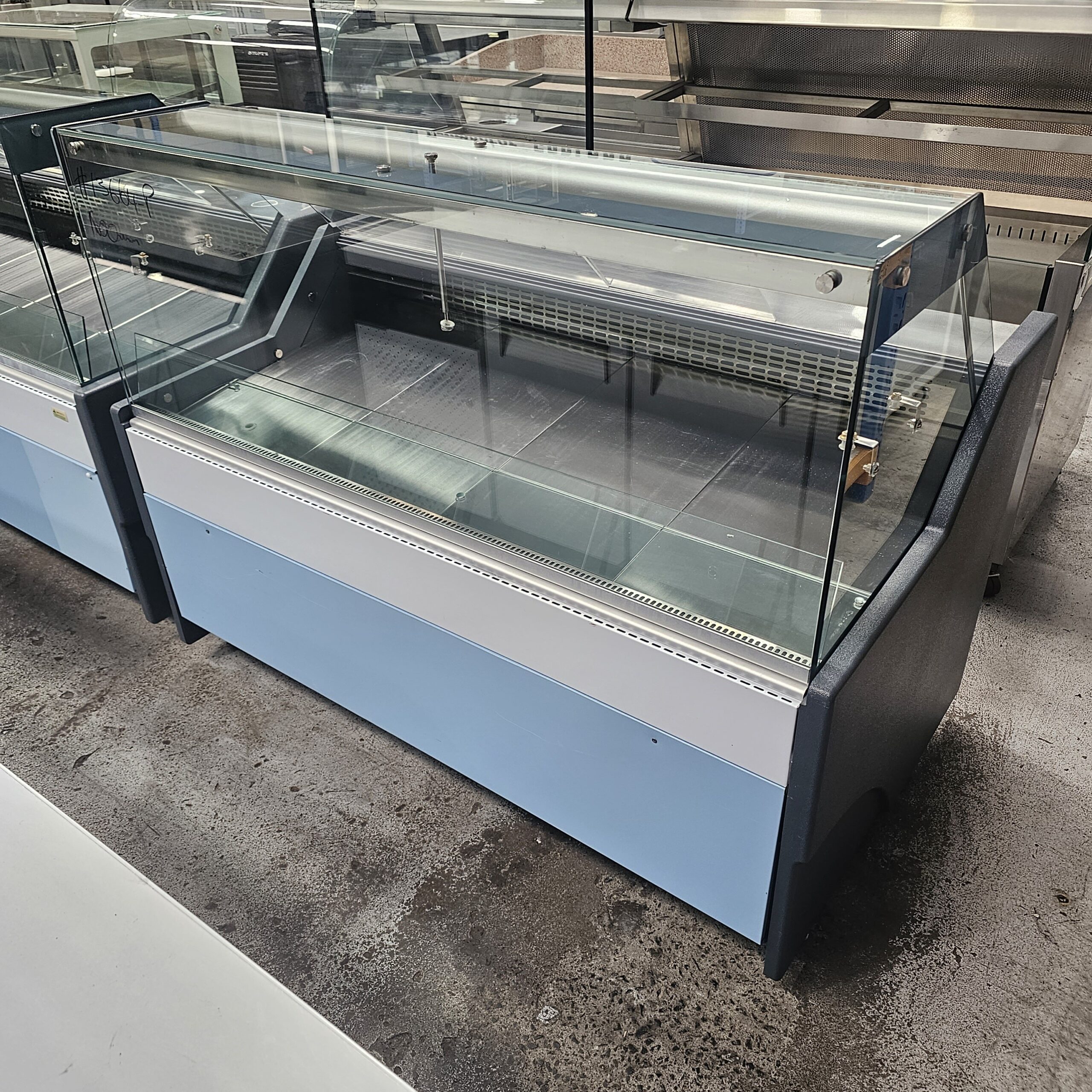 A large, empty refrigerated display case with glass panels and a blue base sits on a concrete floor in a commercial or supermarket setting. MRCE