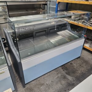 A large, empty refrigerated display case with glass panels on top and sides, situated in a commercial kitchen or warehouse setting with metal shelves and equipment in the background. MRCE
