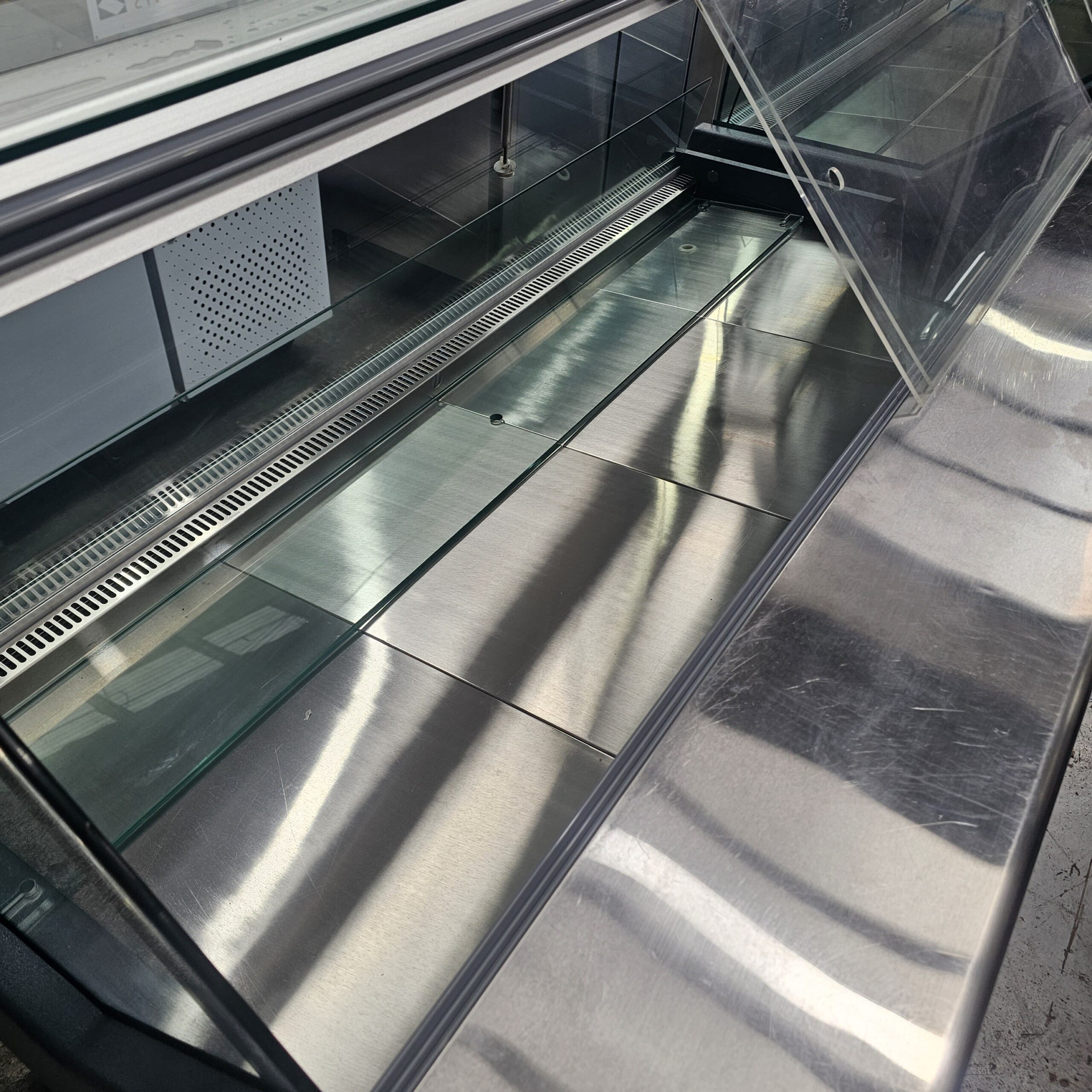 A close-up of an empty stainless steel food display case with the glass door open, showing a clean, reflective surface and no food items inside. MRCE
