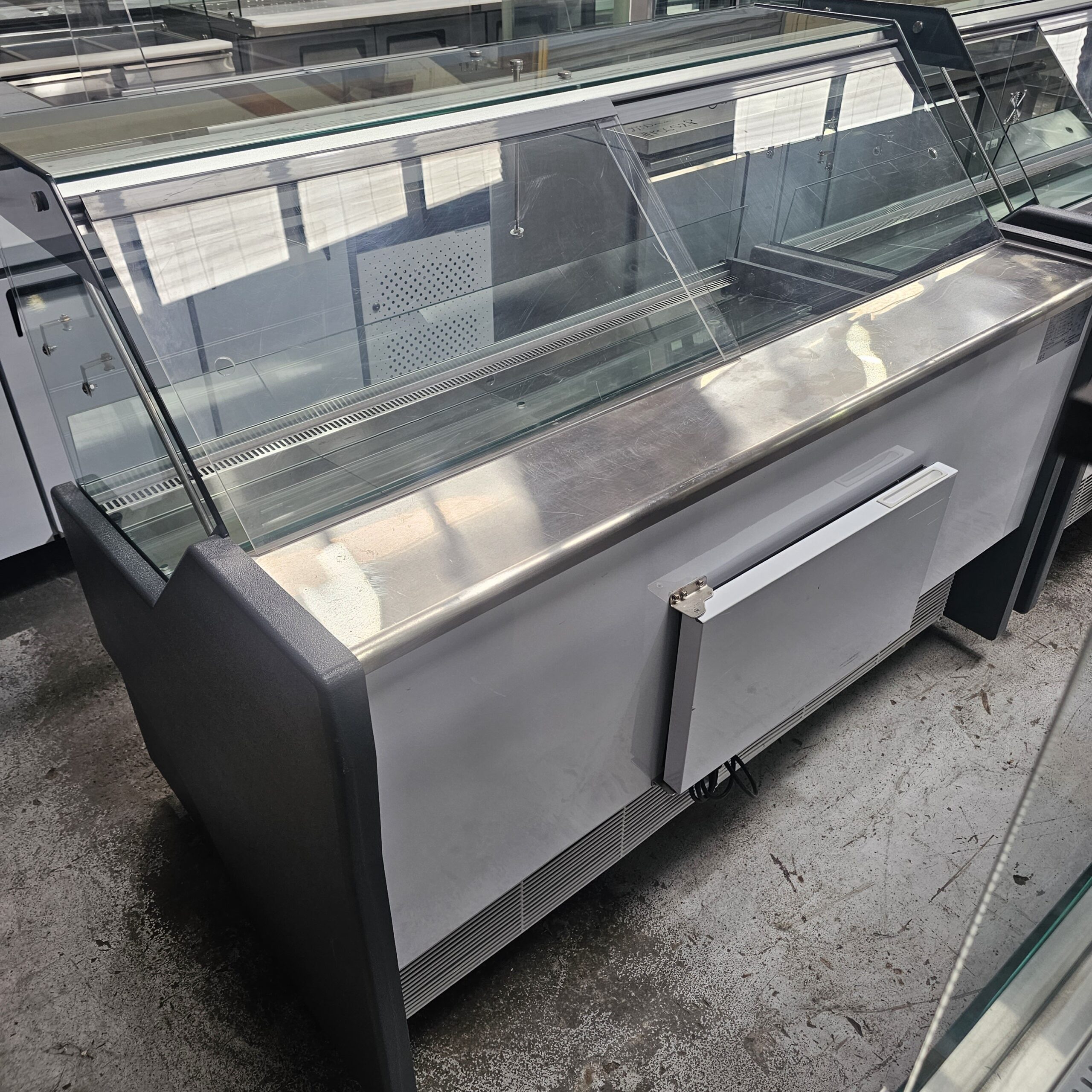 A large, empty glass display case with a stainless steel counter sits indoors on a concrete floor, reflecting light from nearby windows. MRCE