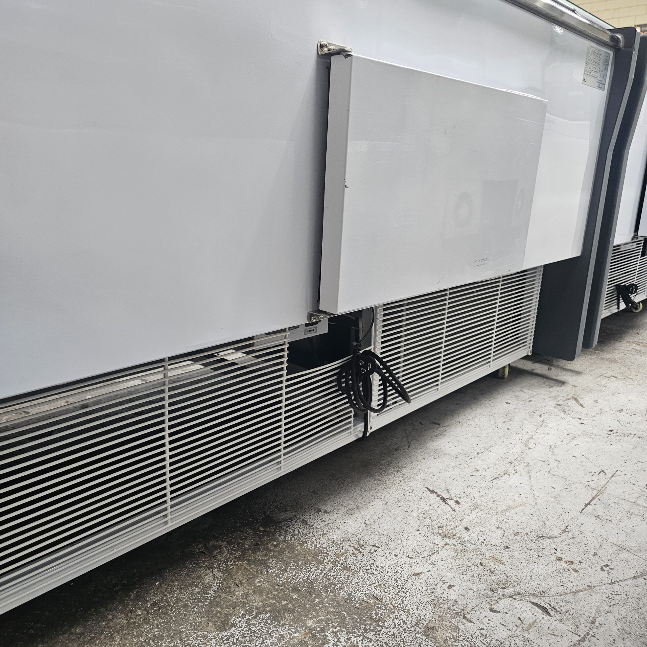 A close-up of the lower side of a large white commercial refrigerator, showing its vented panels, a rectangular access panel, and a black coiled power cord on a concrete floor. MRCE