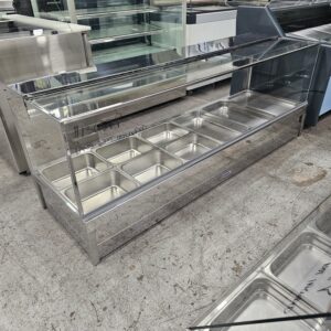 A stainless steel and glass food display case with eight empty metal trays, sitting on a concrete floor in what appears to be a showroom or warehouse. MRCE