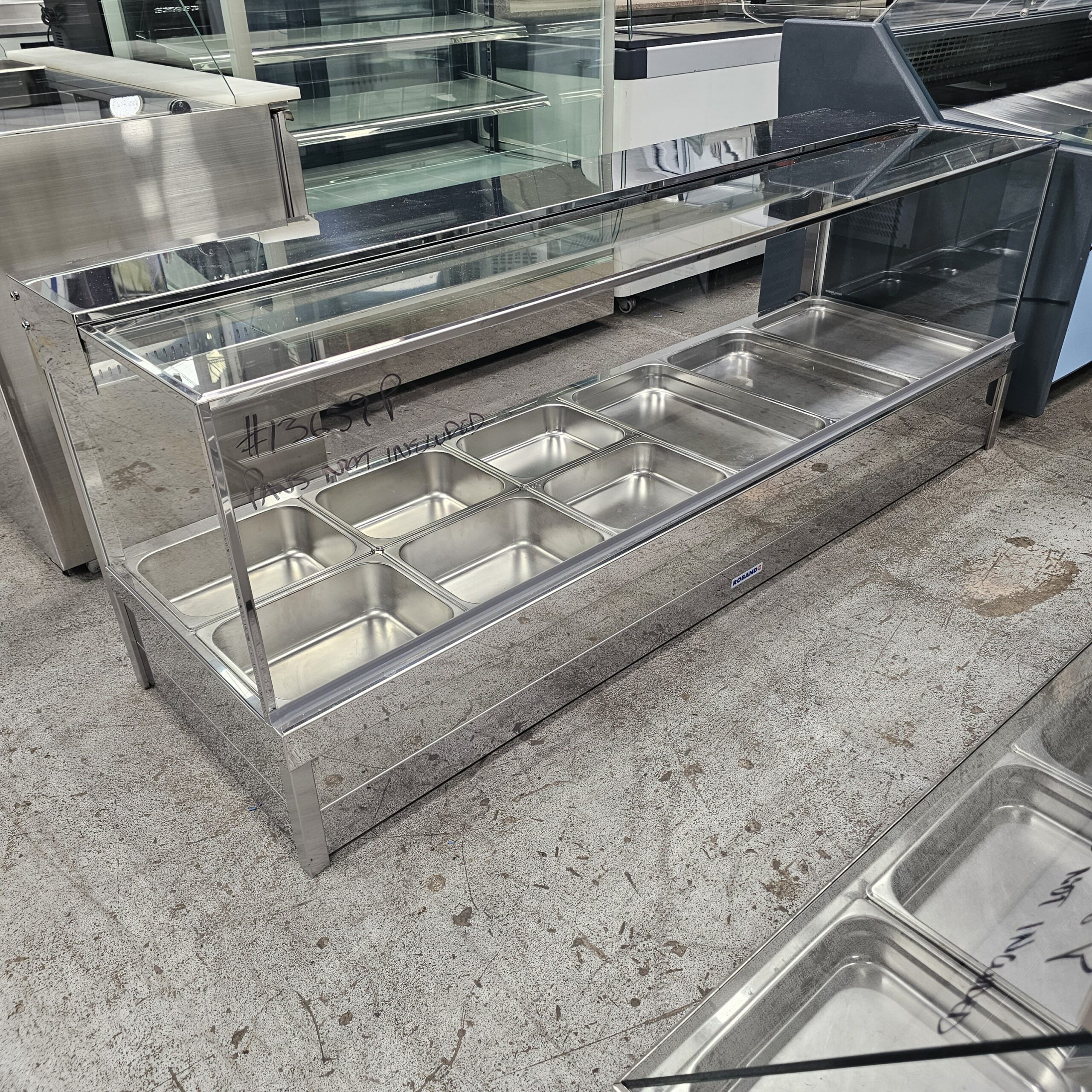 A stainless steel and glass food display case with eight empty metal trays, sitting on a concrete floor in what appears to be a showroom or warehouse. MRCE