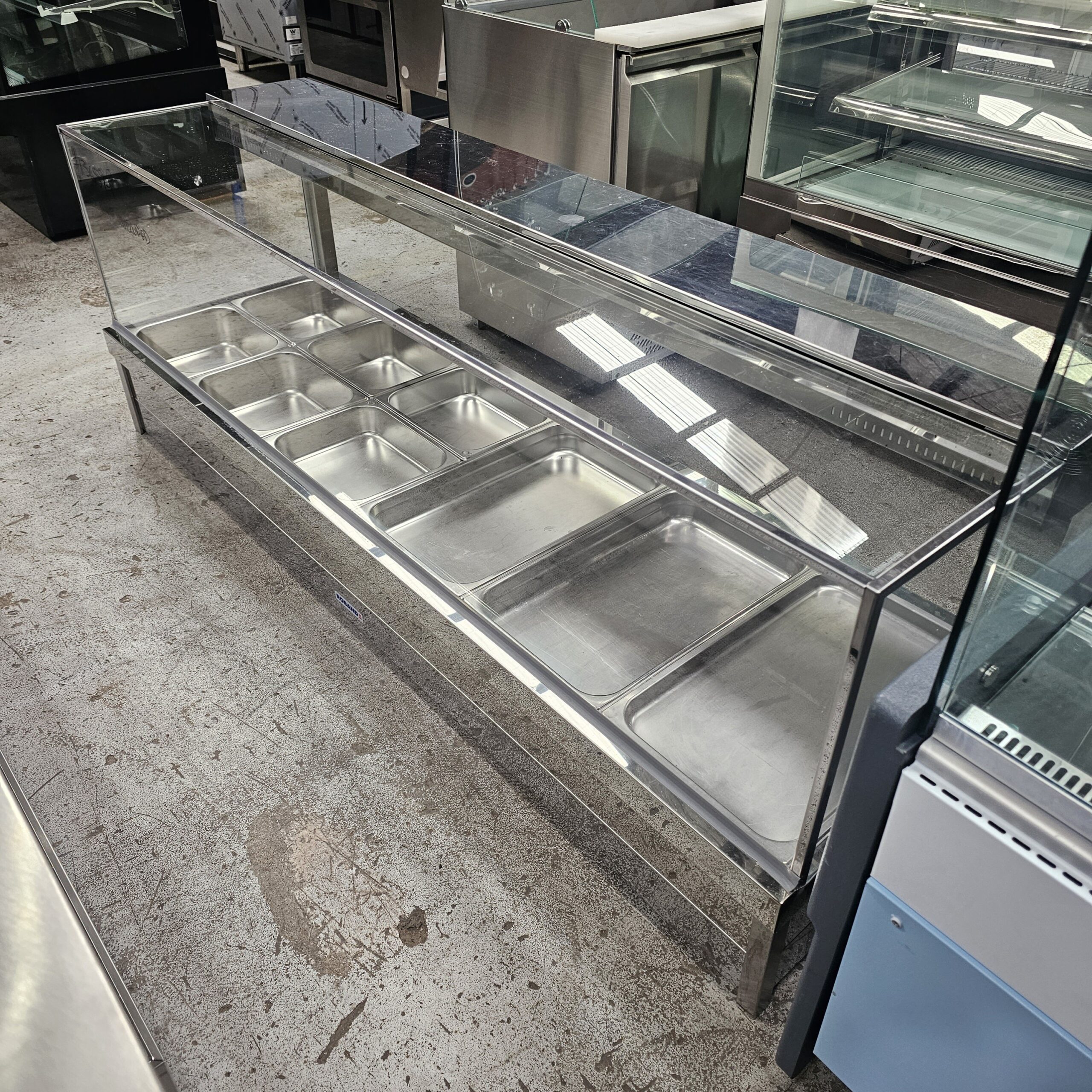 A long, empty stainless steel food display counter with multiple tray compartments and a glass sneeze guard, placed indoors on a concrete floor among other kitchen equipment. MRCE