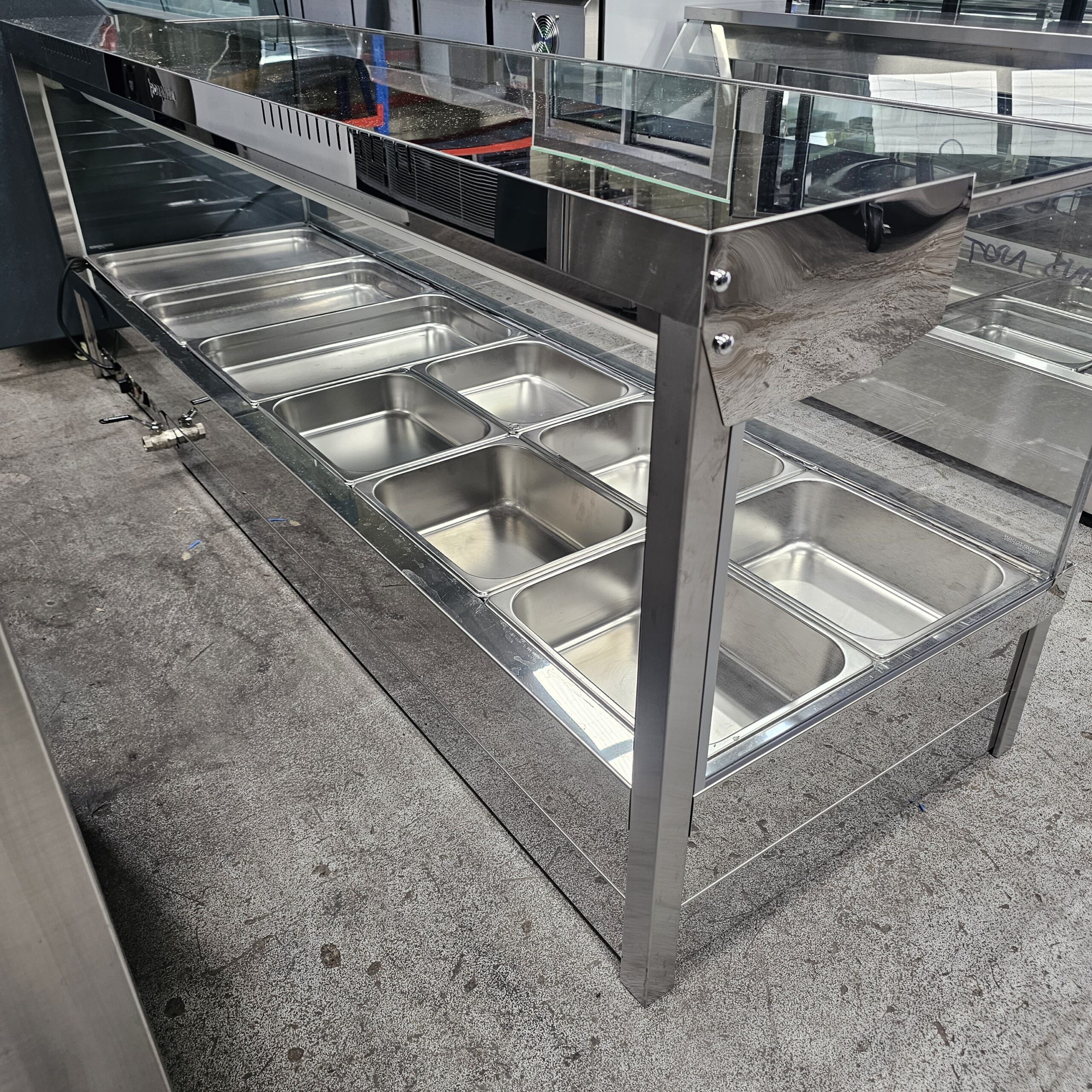 A stainless steel food service counter with eight empty metal trays and a glass sneeze guard, set up on a gray concrete floor in a commercial kitchen or cafeteria. MRCE