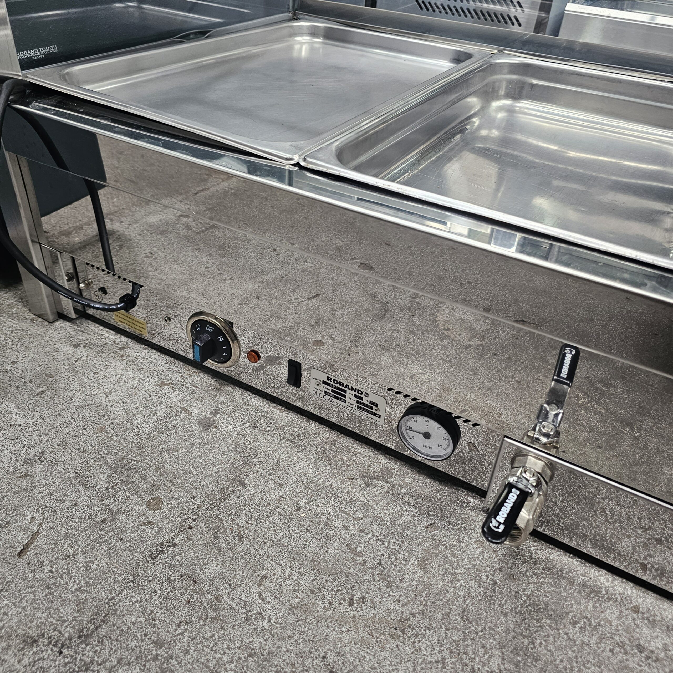 A close-up of a stainless steel food warmer with two covered trays on top, a temperature gauge, a control dial, and a drain valve, all situated on a concrete floor. MRCE