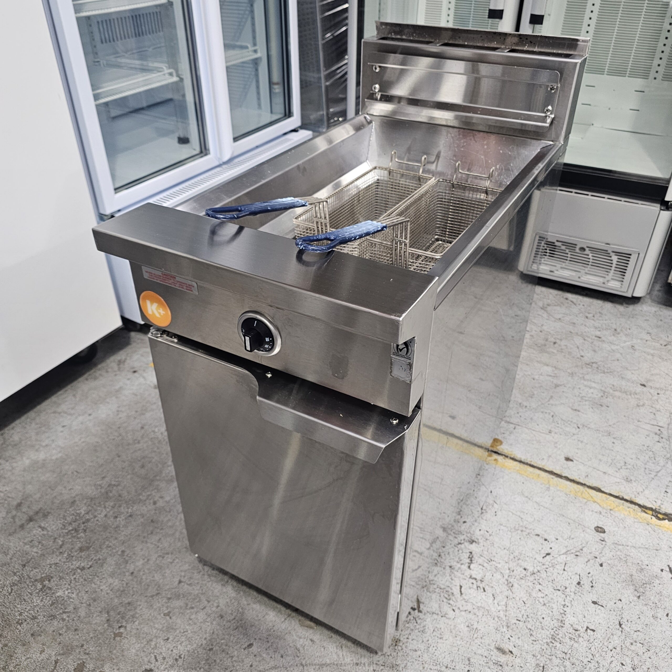 A commercial stainless steel deep fryer with two wire baskets and blue handles, situated in a kitchen or industrial setting with other appliances visible in the background. MRCE