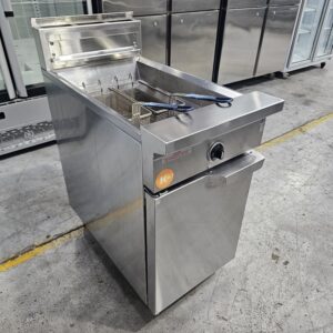 A commercial stainless steel deep fryer with a basket and blue-handled tongs, standing on a concrete floor in an industrial kitchen or storage area. Refrigerators are visible in the background. MRCE