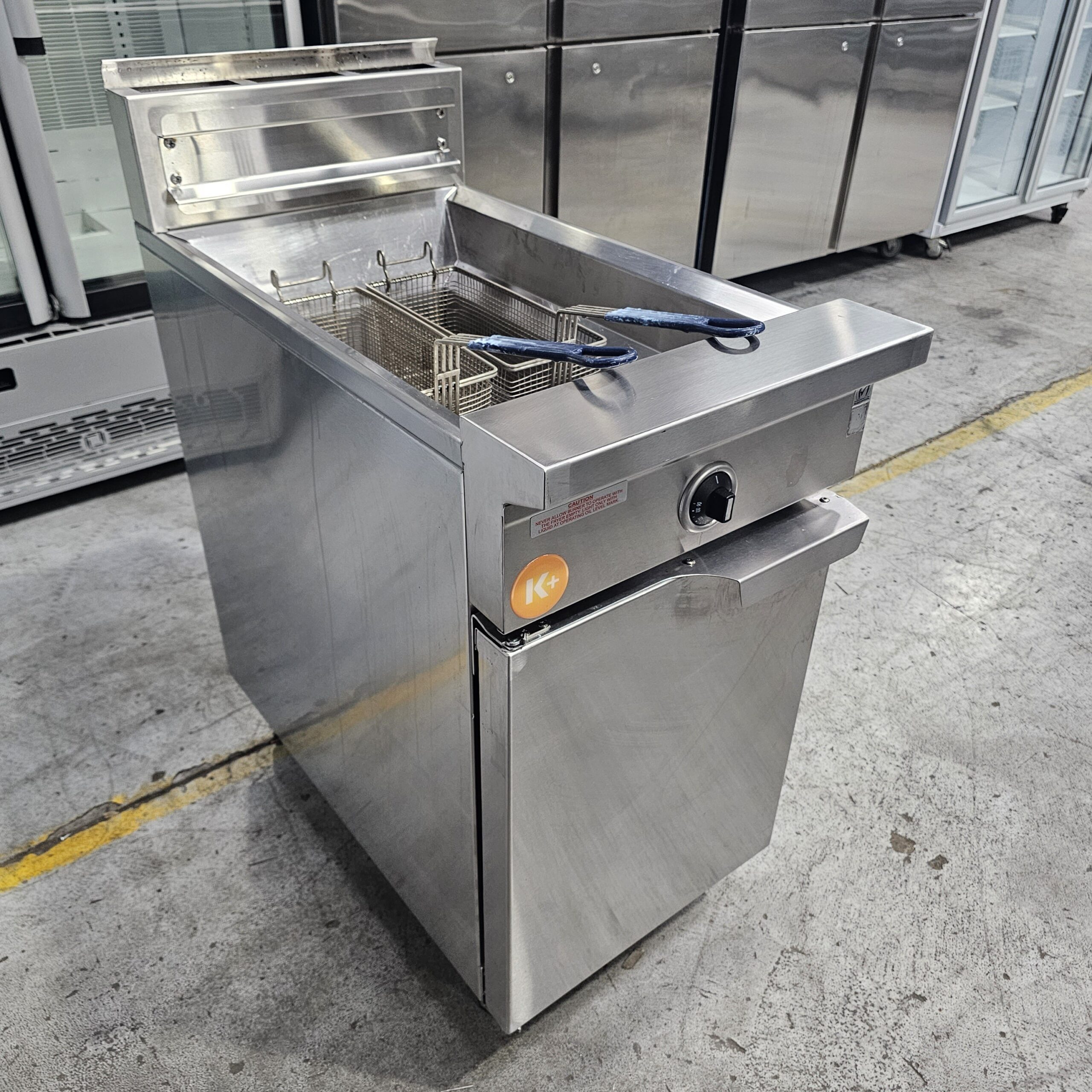 A commercial stainless steel deep fryer with a basket and blue-handled tongs, standing on a concrete floor in an industrial kitchen or storage area. Refrigerators are visible in the background. MRCE