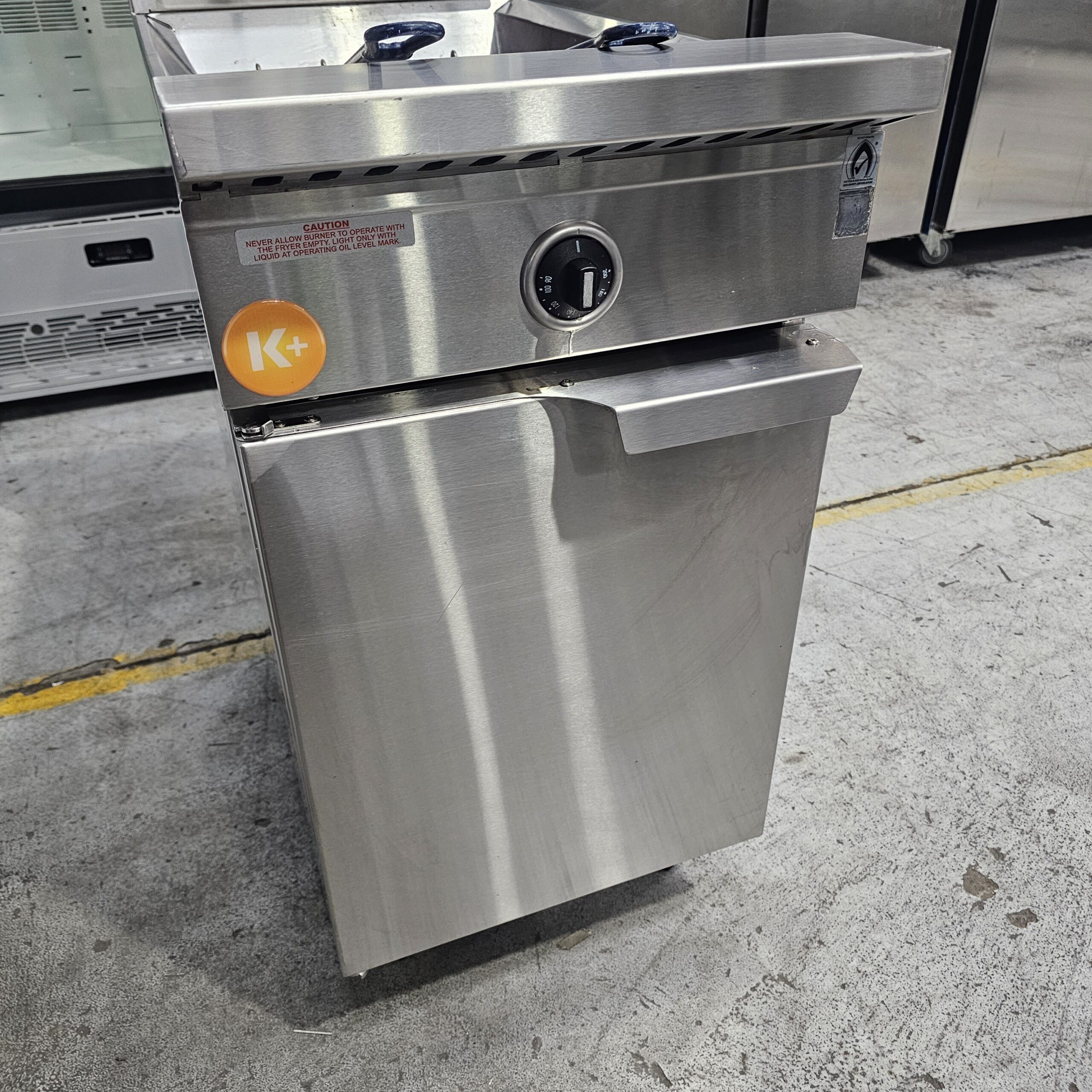 A stainless steel commercial kitchen appliance with a control knob on the front and a Kosher certification sticker, standing on a concrete floor near a yellow line. MRCE