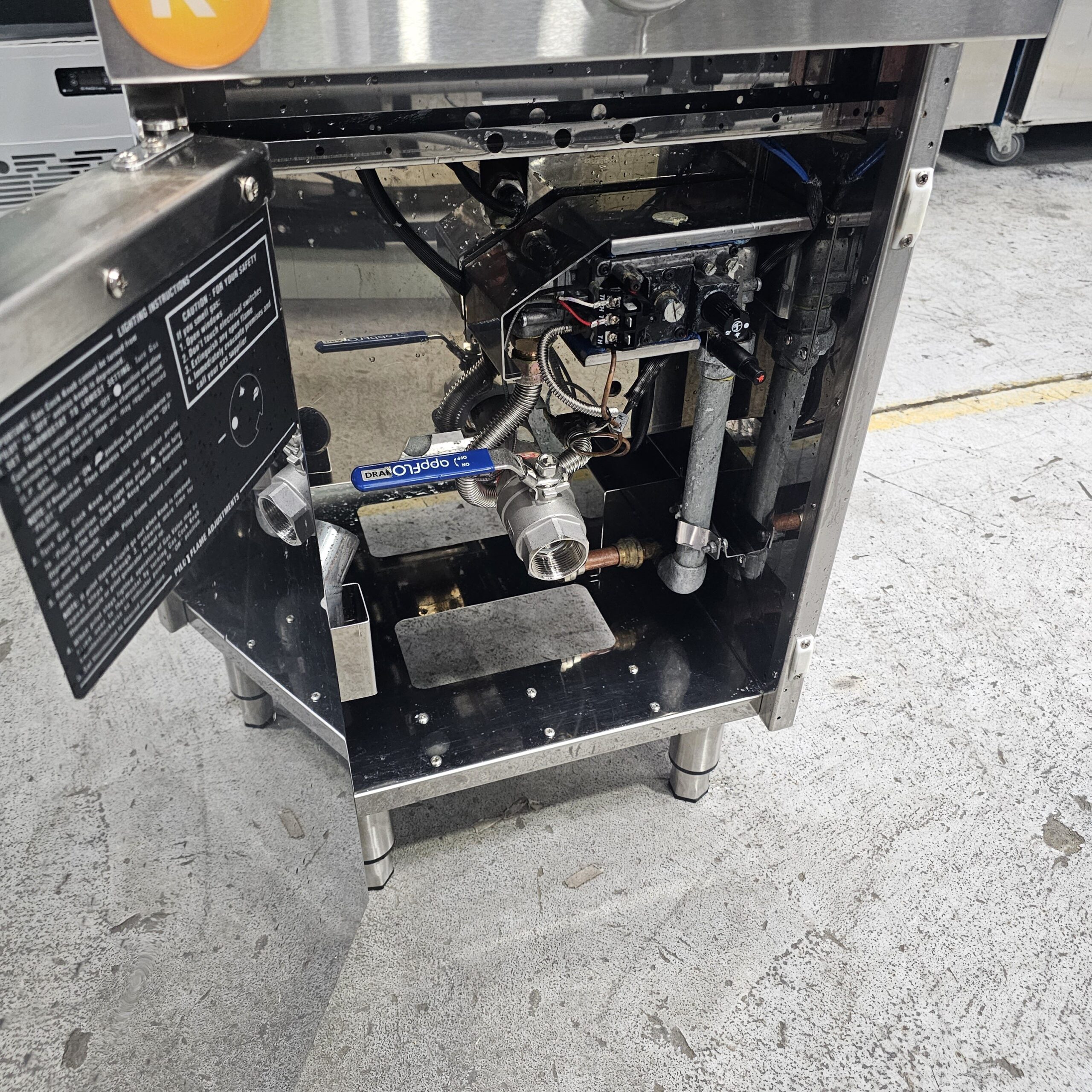 The image shows the open lower compartment of an industrial machine, revealing complex internal components, pipes, and wires. The machine is situated on a concrete floor with a yellow line visible nearby. MRCE