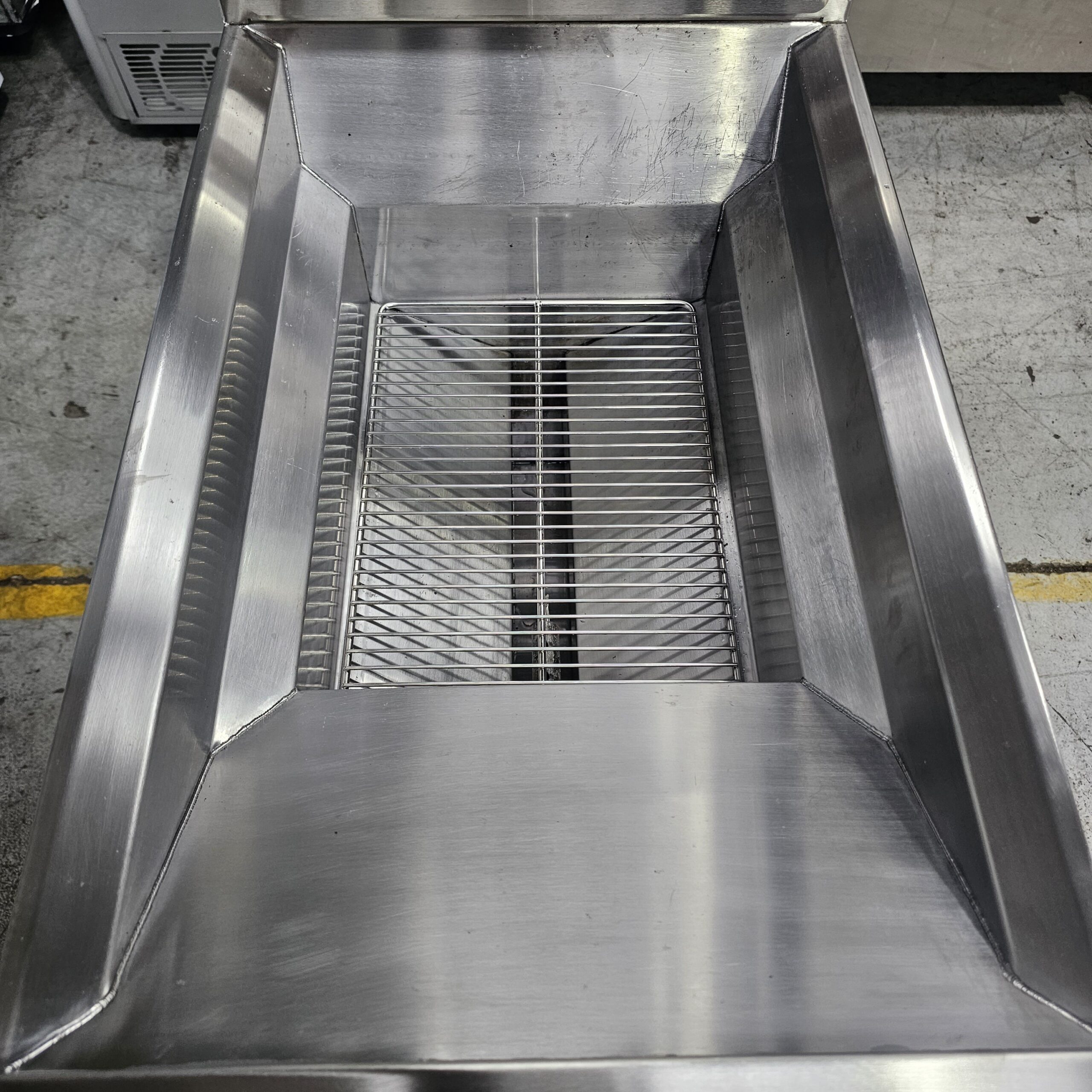 A close-up view of a stainless steel commercial kitchen sink or basin with a metal grate at the bottom, set on a concrete floor with a yellow line in the background. MRCE